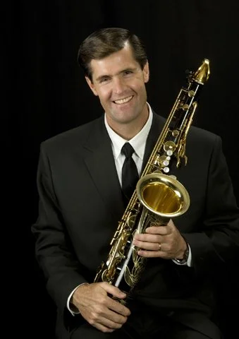 Dr. Mark Watkins: Smiling man in a black suit holding a gold alto saxophone against a black background.