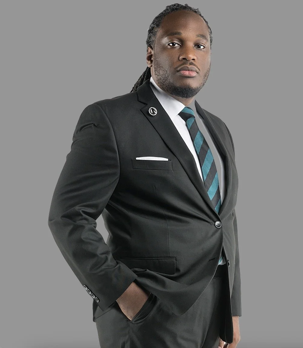 DR. SYDNEY FREEMAN JR.
A man with dreadlocks wearing a black suit, white shirt, and striped tie against a gray background.