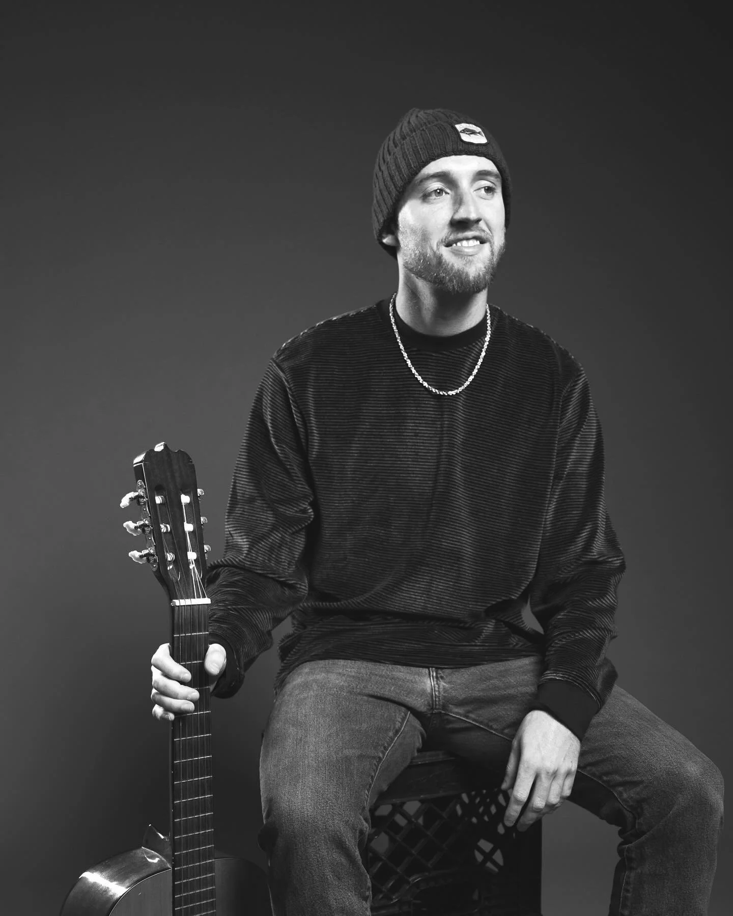 A young man sitting with a guitar on his lap, wearing a beanie, a chain necklace, a long-sleeve shirt, and jeans, smiling against a plain dark background.