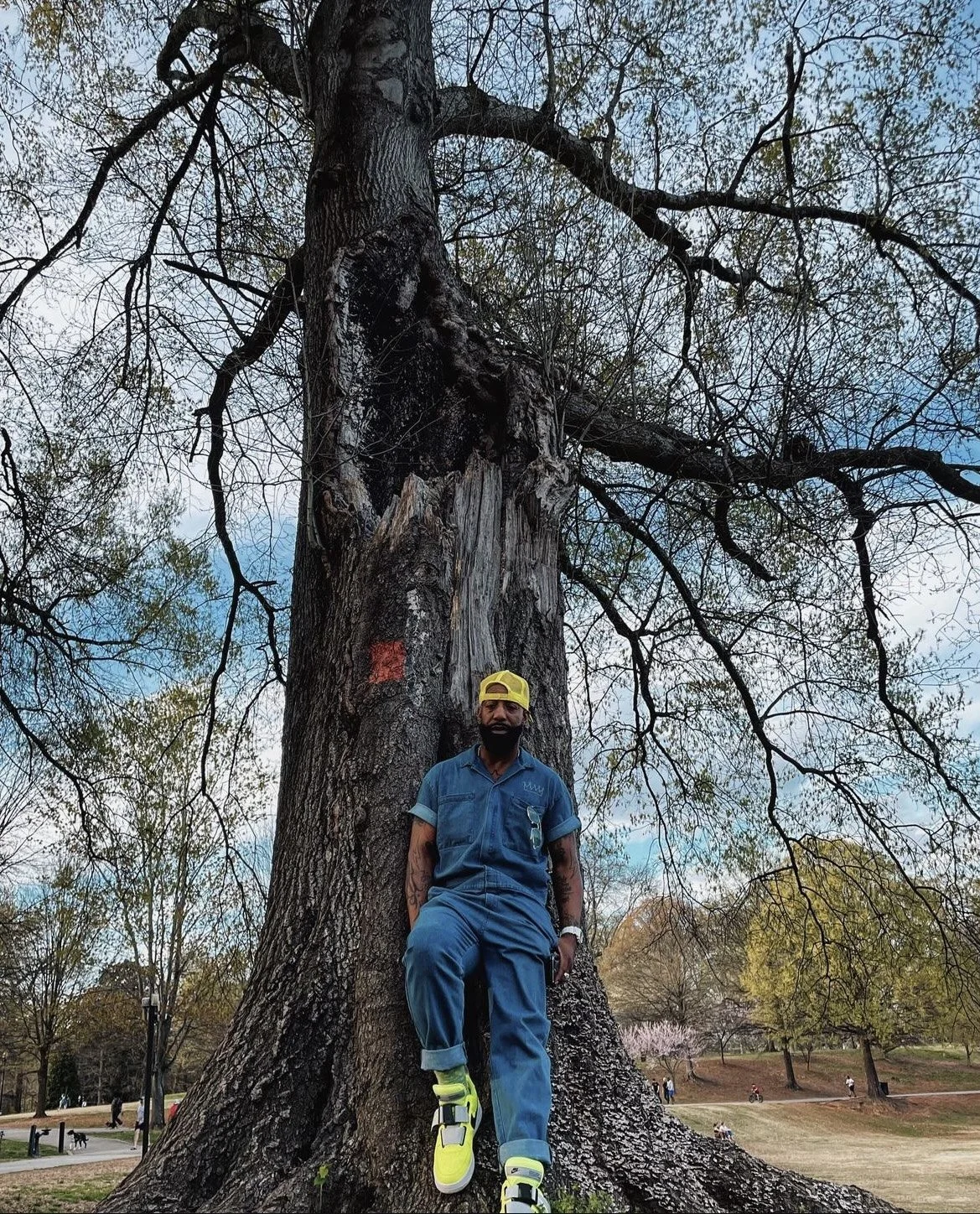 A man with a beard, wearing a yellow cap, blue jumpsuit, and neon yellow sneakers, stands by a large tree in a park.