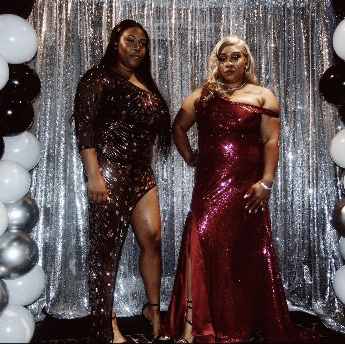 Two women in glamorous red sequined dresses standing in front of a silver sequin backdrop with black and white balloons on the sides, at an event or celebration.