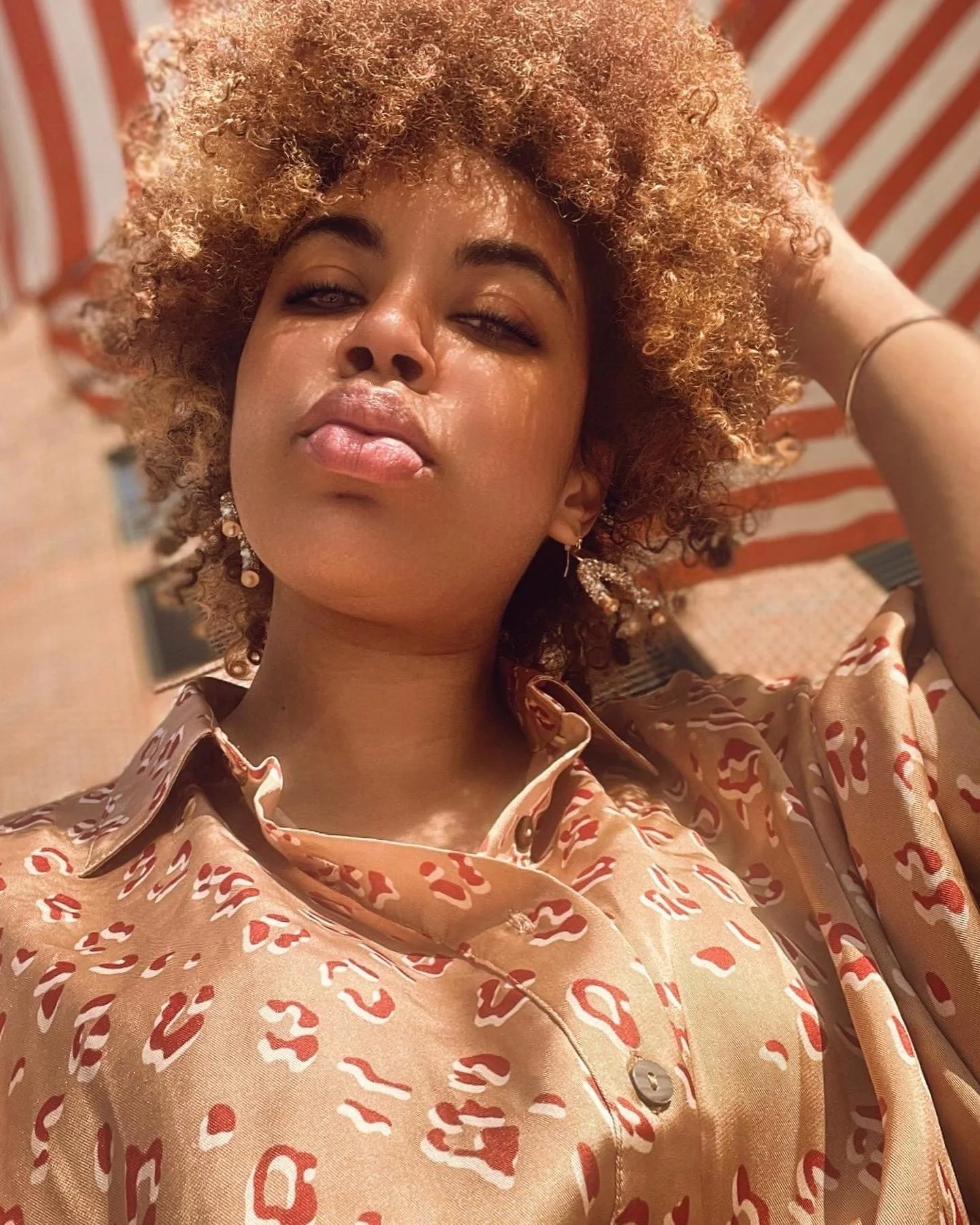 A woman with natural curly hair, wearing earrings and a patterned shirt, takes a selfie outdoors with a red and white striped canopy in the background.