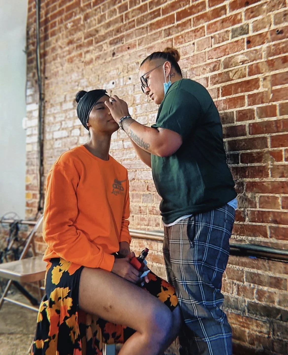 A woman getting a tattoo on her thigh while a tattoo artist works on her leg, set against an exposed brick wall.