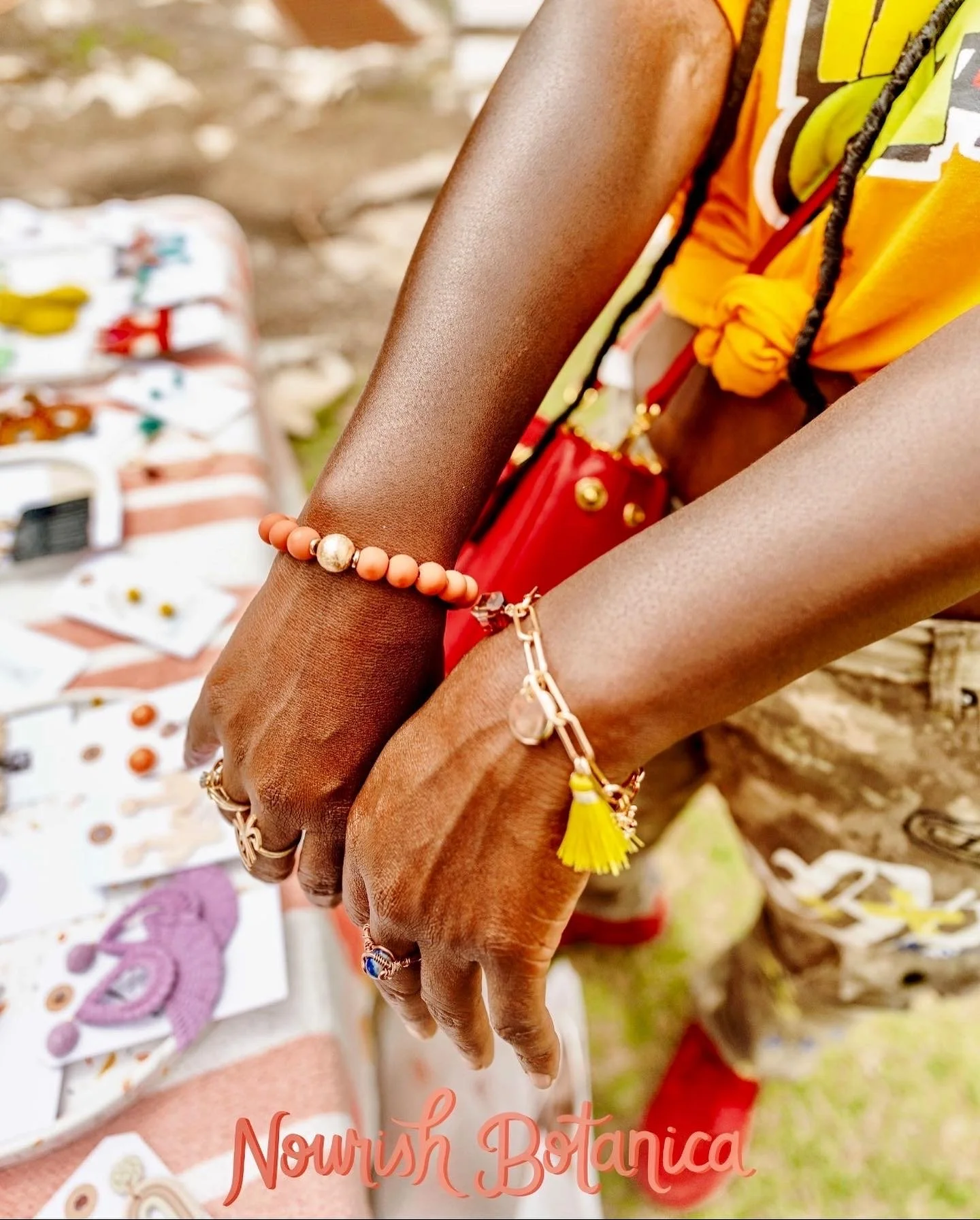 Two hands wearing jewelry with a background of jewelry and accessories on display, and the words 'Nourish Botanica' at the bottom.