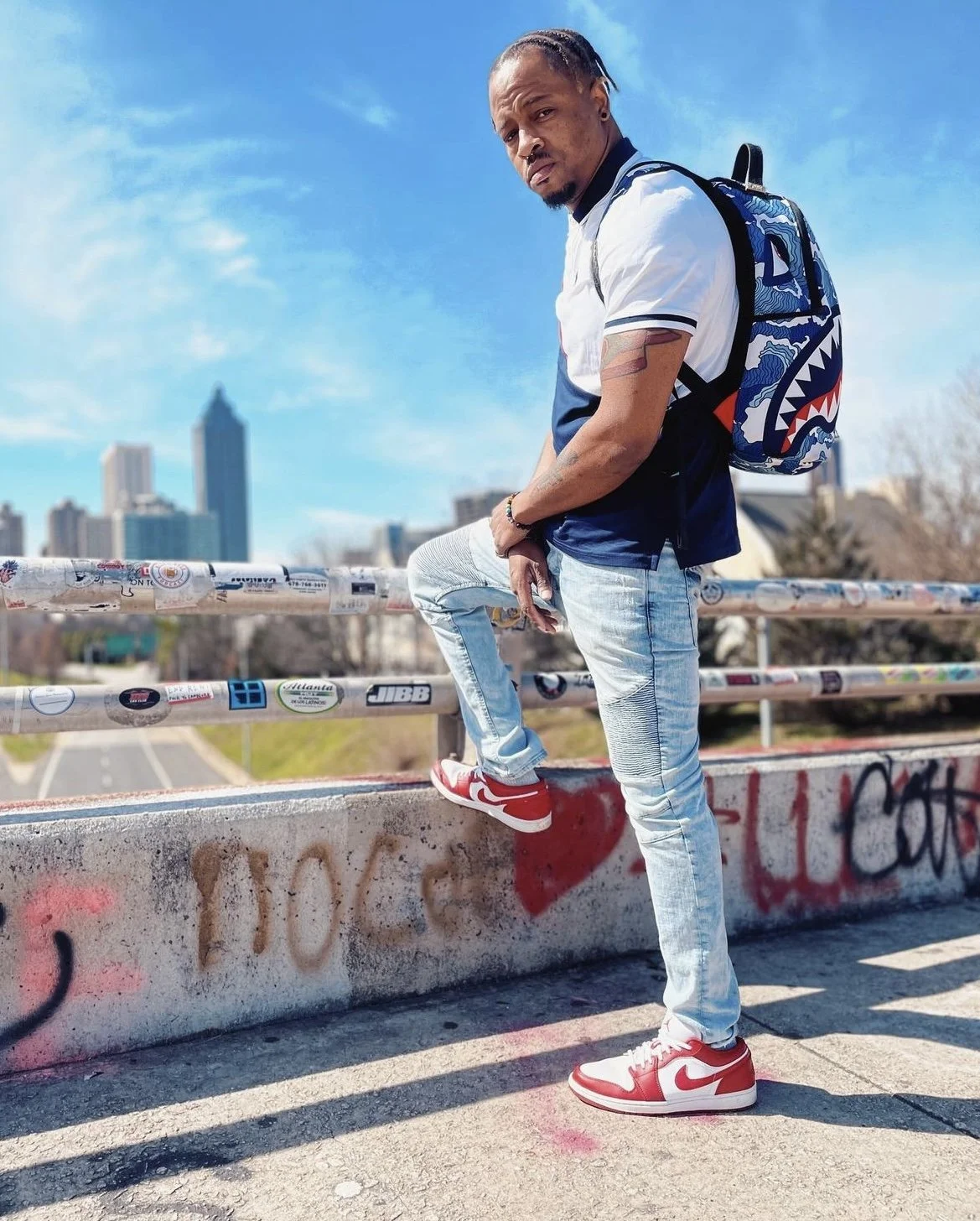 A man with braided hair and tattoos wearing a white and blue shirt, light blue jeans, and red and white Nike sneakers, standing on a graffiti-covered concrete ledge with the Atlanta skyline in the background on a sunny day.