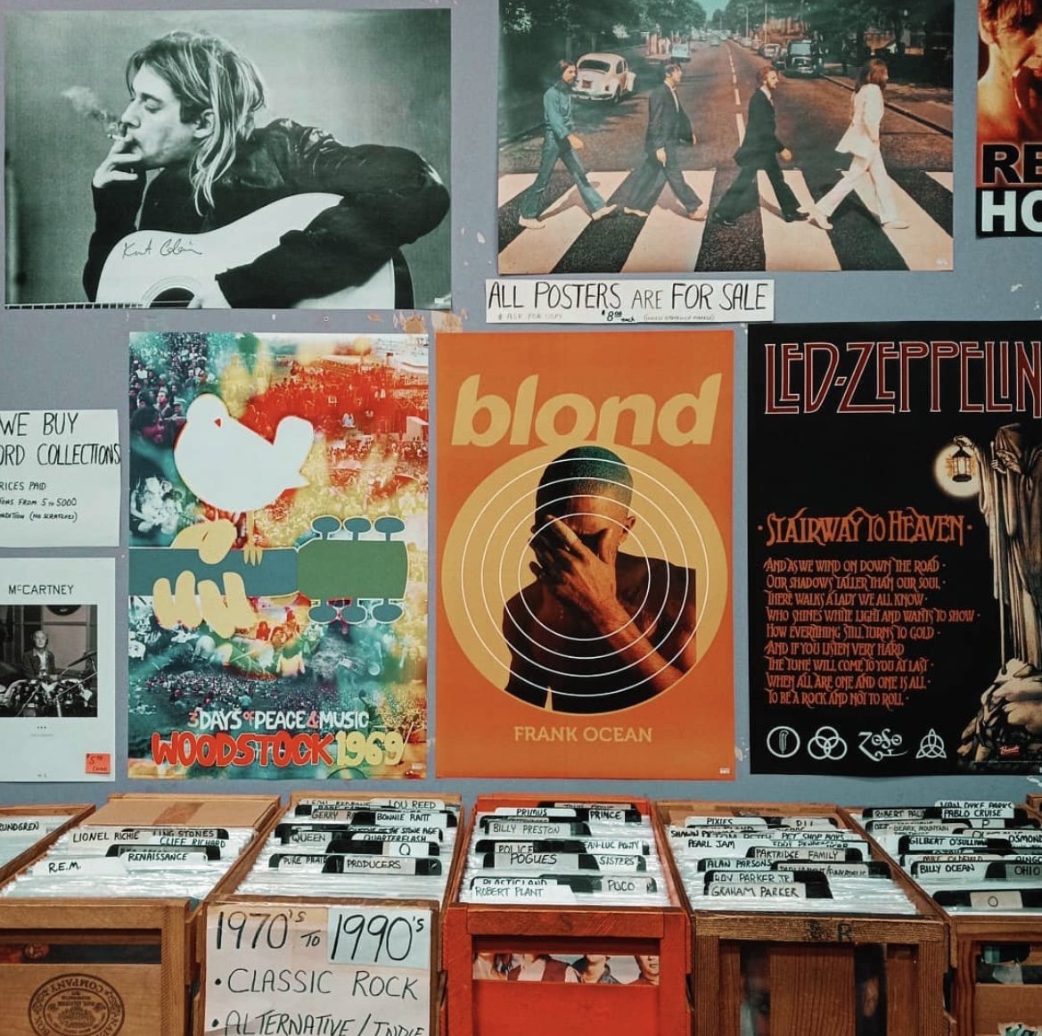 A wall with vintage posters including a woman smoking and playing guitar, The Beatles Abbey Road album cover, a colorful Woodstock 1969 poster, a Blond album cover by Frank Ocean, and a Led Zeppelin Stairway to Heaven poster. Below, there are bins of