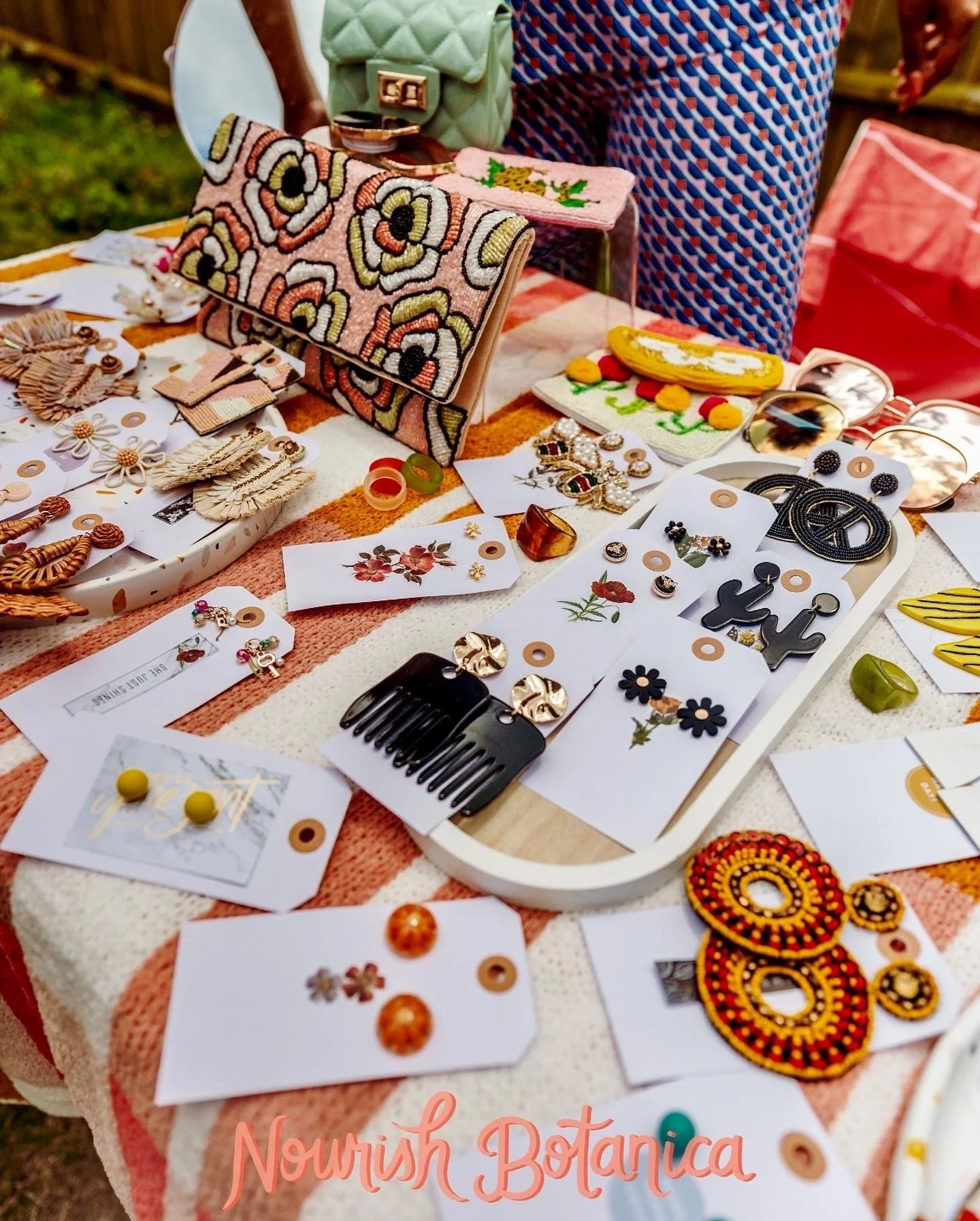 A table at an outdoor market displaying various colorful accessories, including earrings, hair combs, and embroidered patches. A quilted purse with a floral design is prominently displayed, along with another small purse and various jewelry pieces, some on white cards.