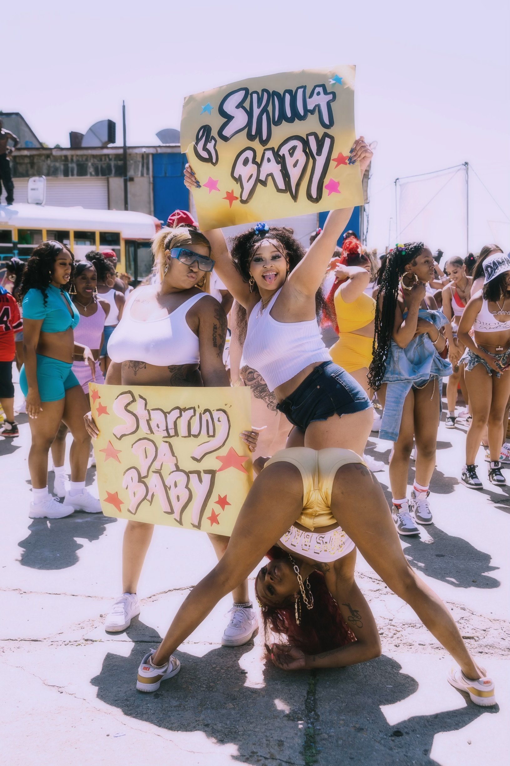 Group of women at a daytime outdoor event, with two women in center holding signs that read 'Starring BABY 2' and a third woman performing an acrobatic pose, all dressed in casual summer clothing.