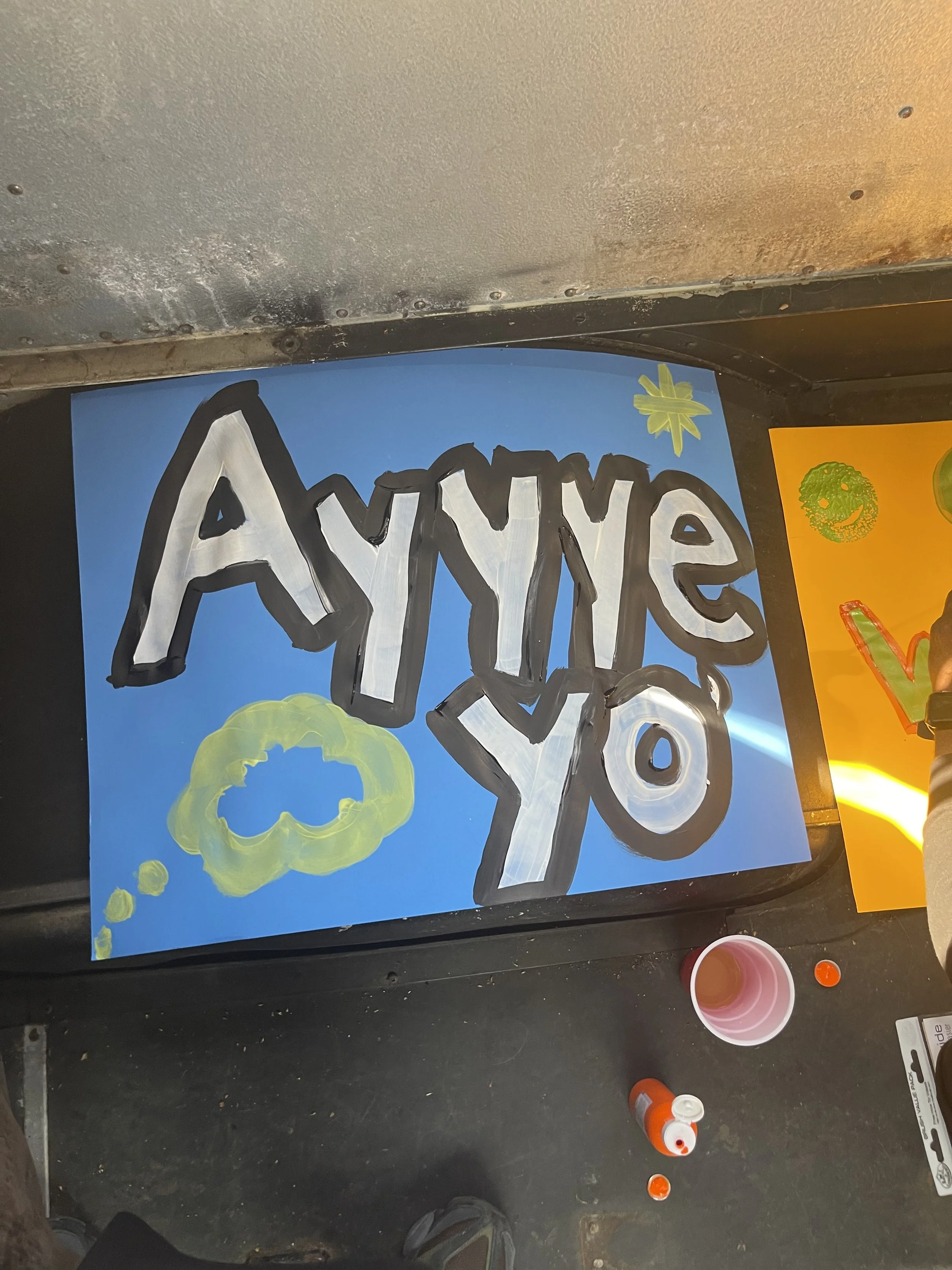A colorful handmade sign with the words "Ayyye Yo" in large, stylized letters, painted in white with black outlines on a blue background. There are decorative elements including a yellow cloud shape and a small green star.