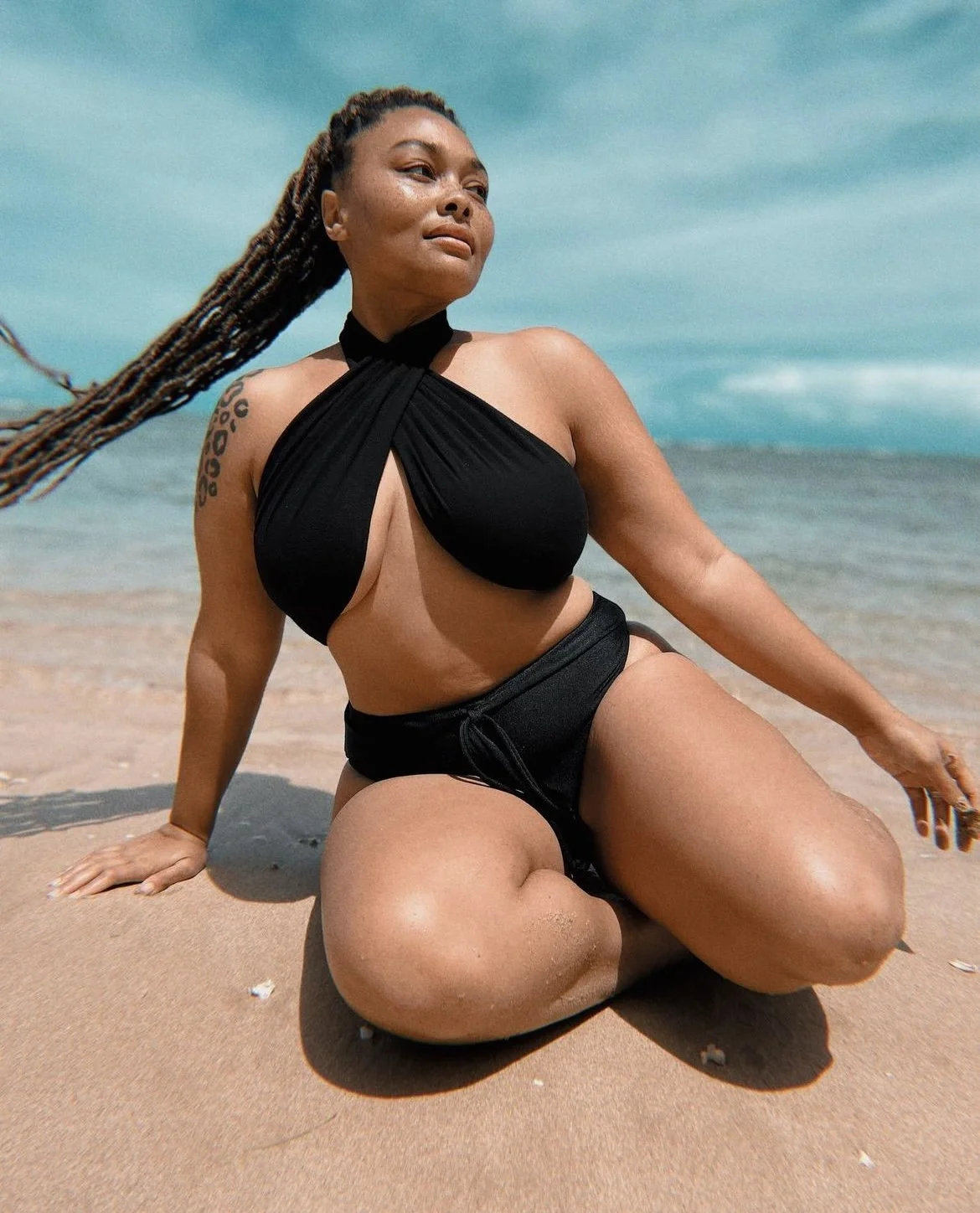 A woman with long braids kneeling on a sandy beach, wearing a black swimsuit with a cutout design, with the ocean and blue sky in the background.