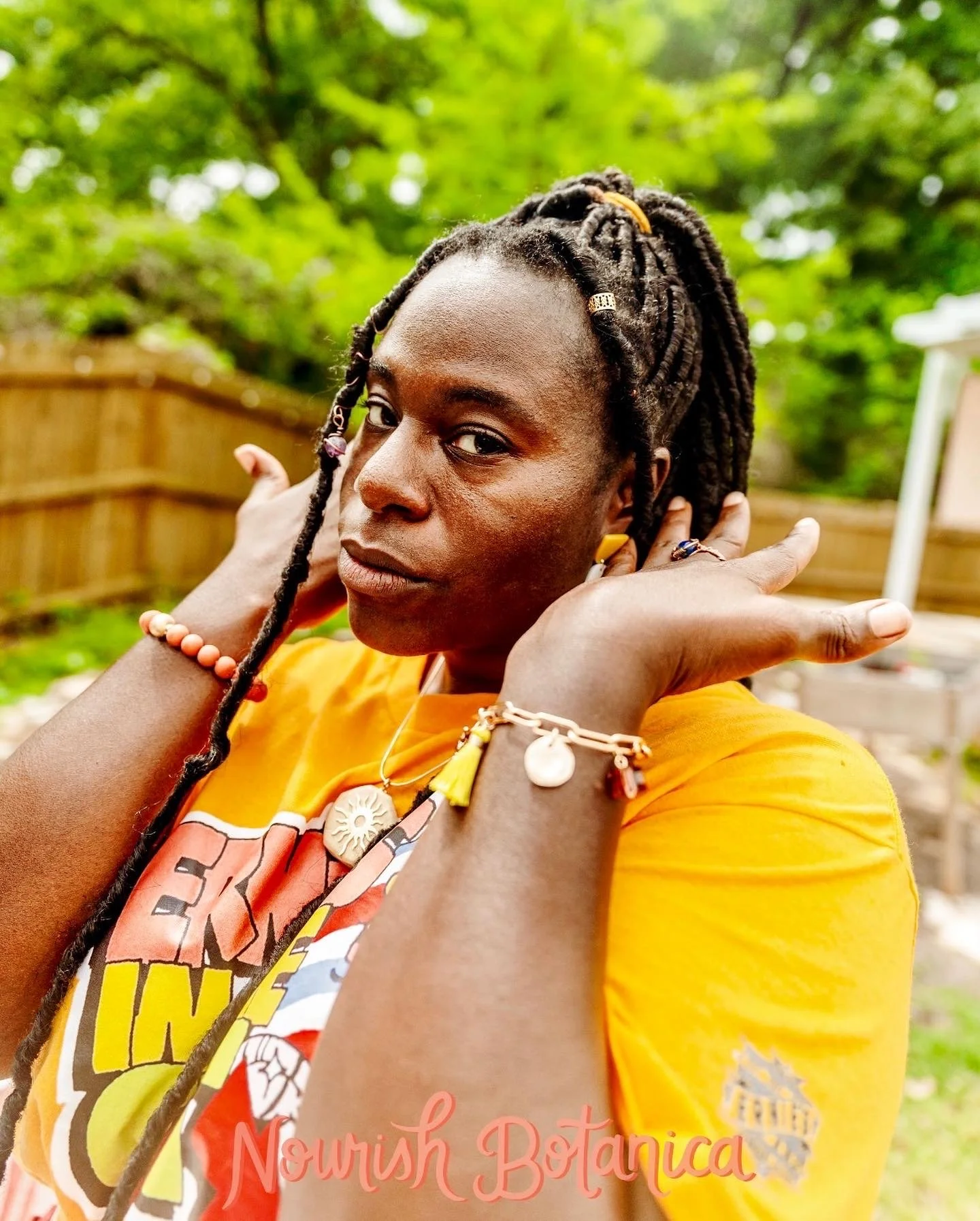 A woman with braided hair and jewelry is posing outdoors in front of a wooden fence and green trees, wearing a yellow shirt. She holds her hands near her face and looks at the camera with a serious expression.