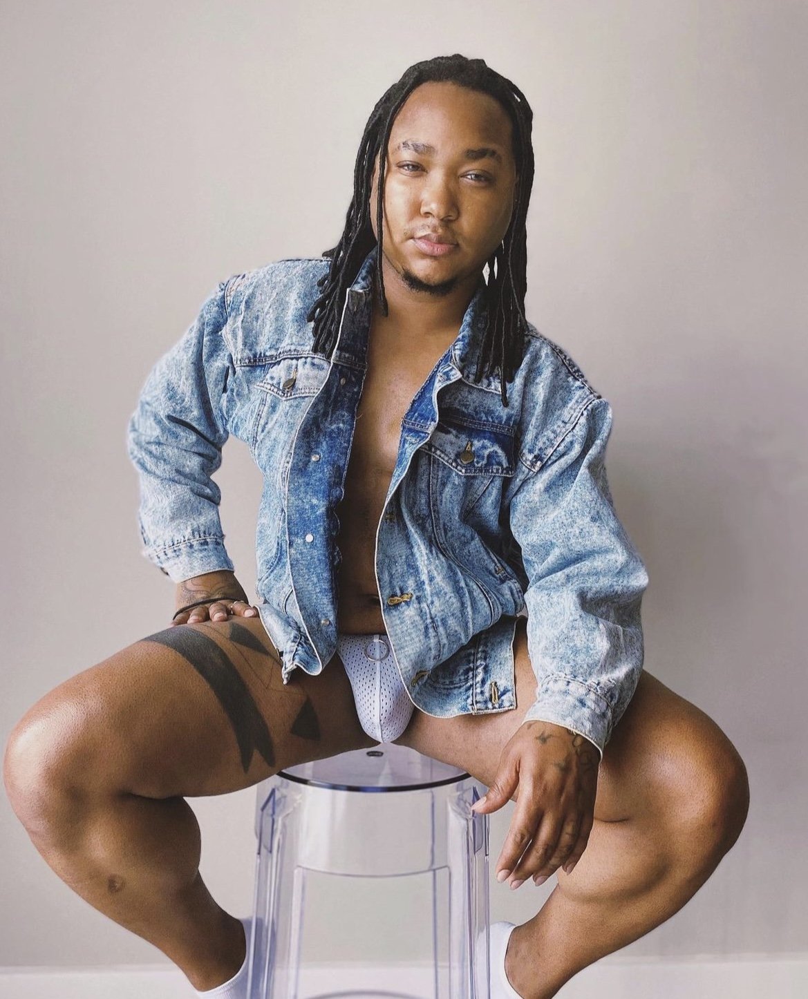 A person with long braided hair wearing a denim jacket and underwear is sitting on a clear stool against a plain background.