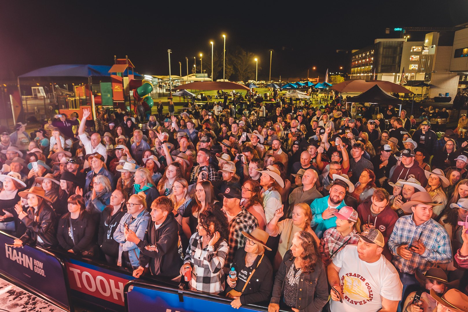 Crowd of people gathered outdoors at night at Sunset Bash Country Music Festival 2024 , some wearing cowboy hats, at a lively event with buildings and a playground in the background.