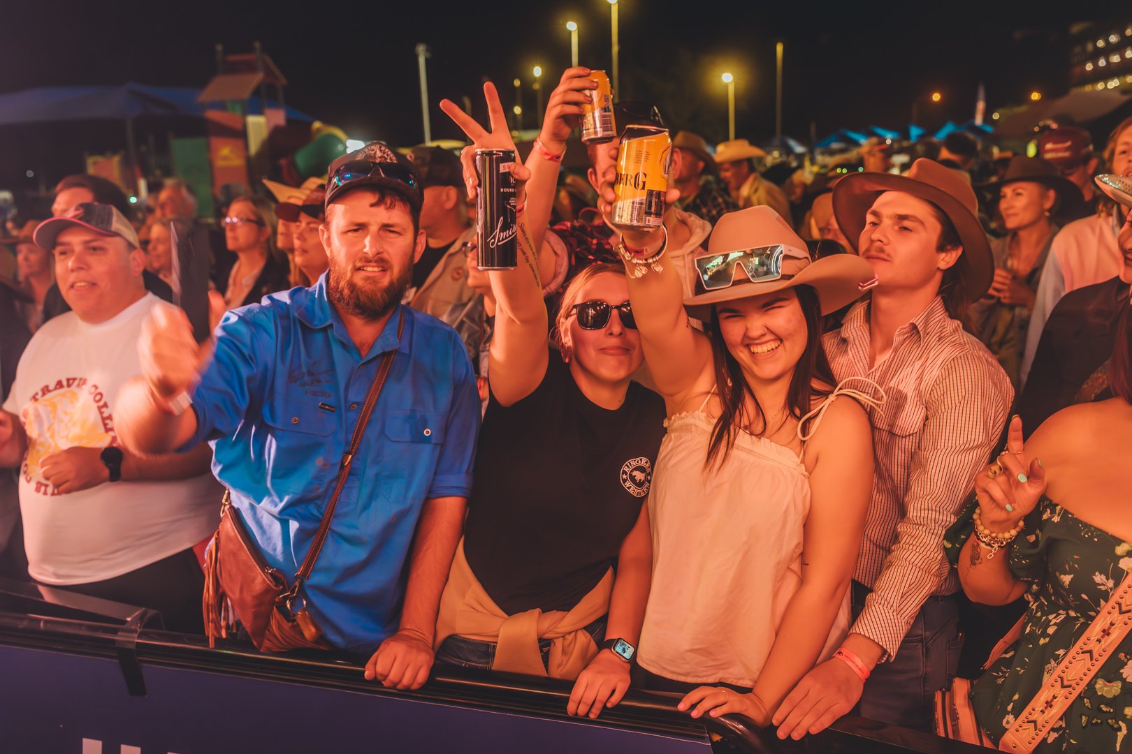 Group of people at Sunset Bash Country Music Festival 2024 enjoying drinks and celebrating, wearing casual and cowboy attire, some with hats and sunglasses, at night with lights overhead.