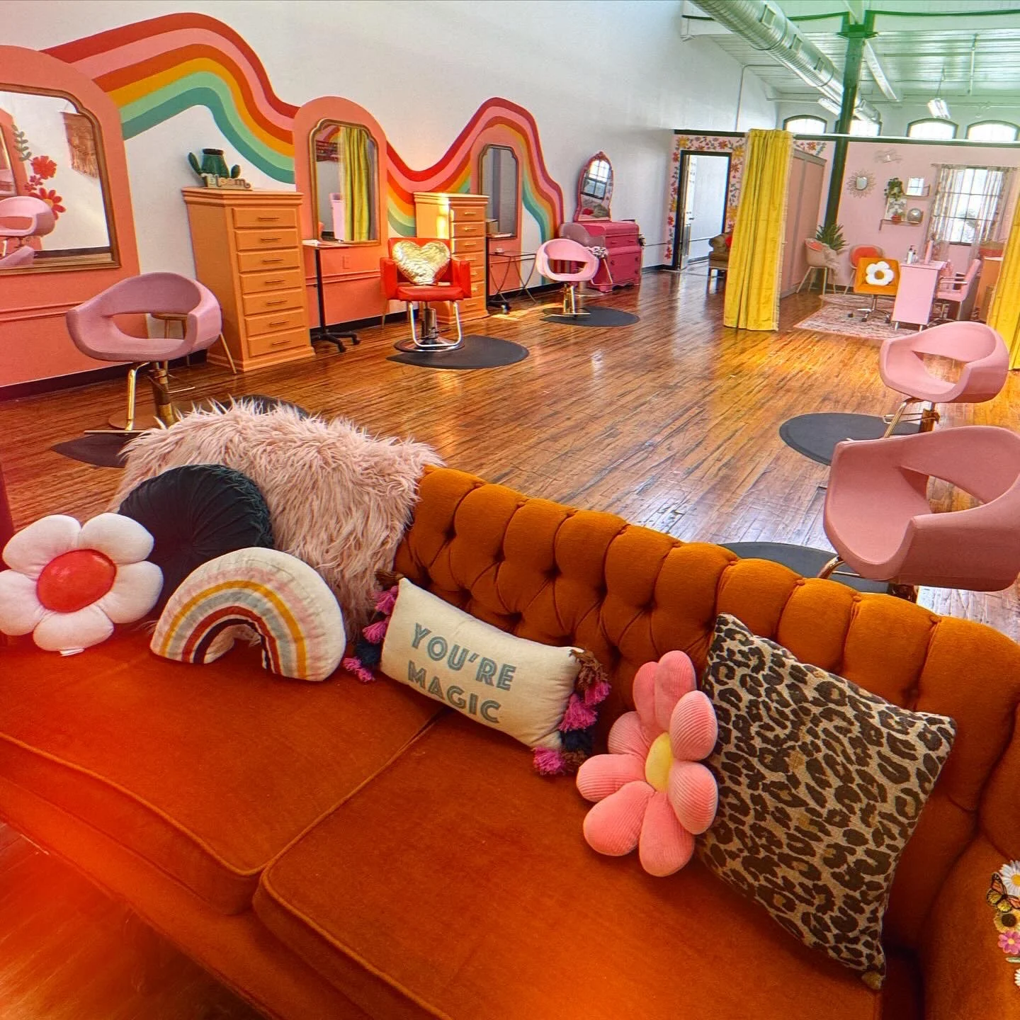 A colorful and retro-style hair salon with pink, orange, and rainbow decor. There's a row of vintage pink salon chairs with black mats, and various furniture pieces in bright colors. The space features a mix of playful cushions, a fuzzy blanket, and decorative pillows, including one with the text 'You're Magic'. The floors are hardwood, and the walls are decorated with rainbow and floral accents.