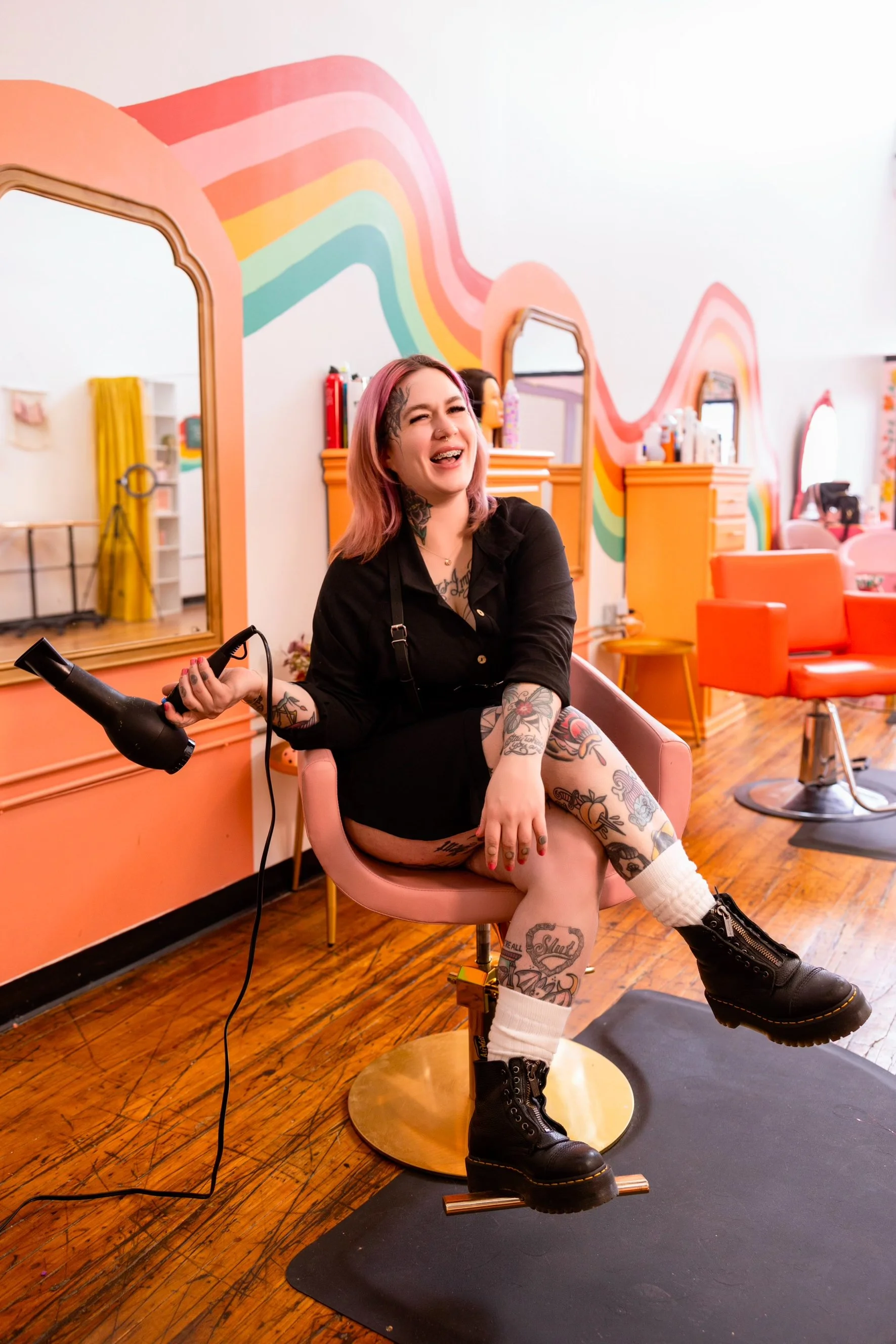 A woman with tattoos and pink hair sitting in a salon chair, laughing, holding a hairdryer. The salon has a vibrant, colorful rainbow mural on the wall and orange furniture.