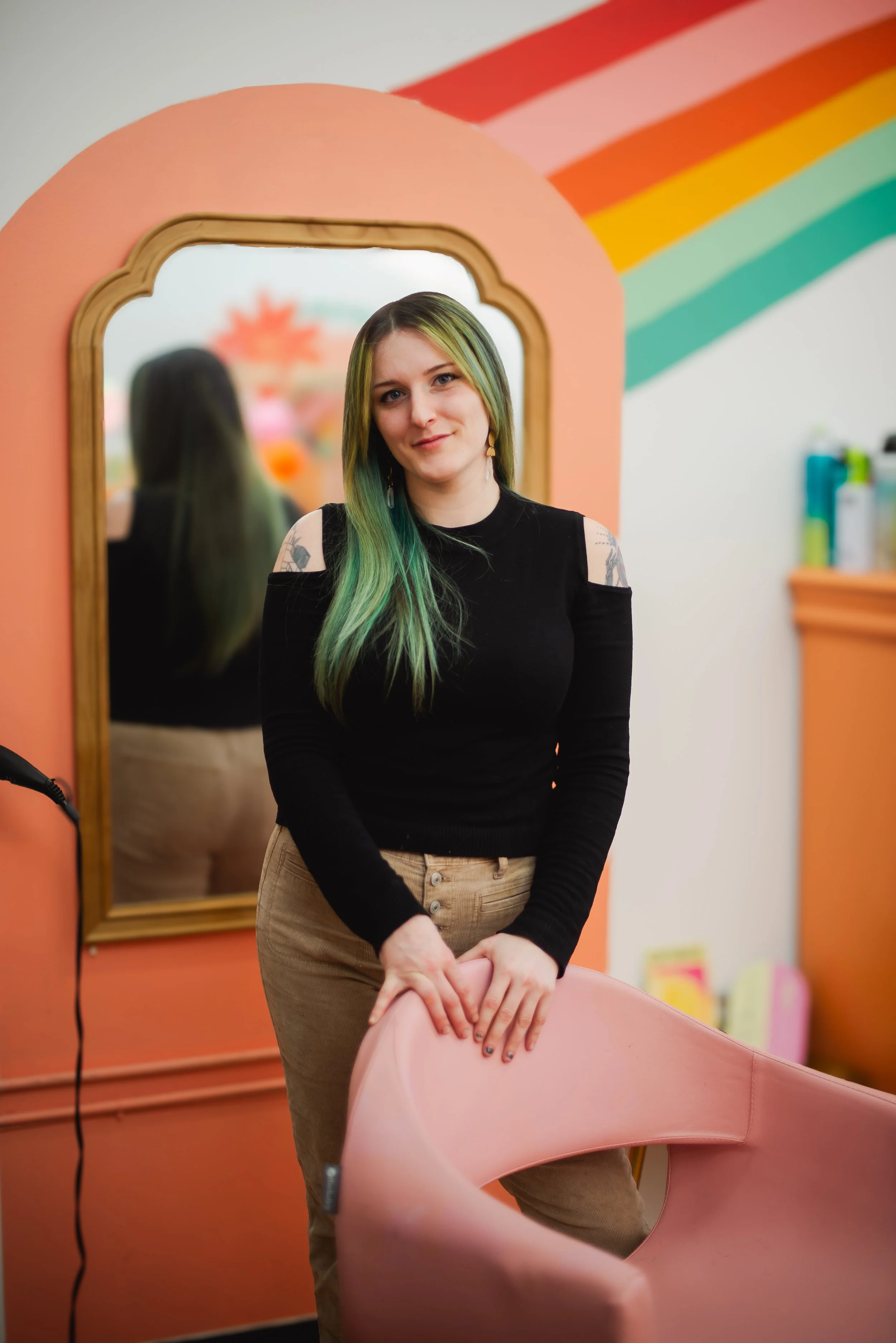 A woman with green hair and tattoos standing in a colorful room with a mirror and rainbow wall art, leaning on a pink chair.
