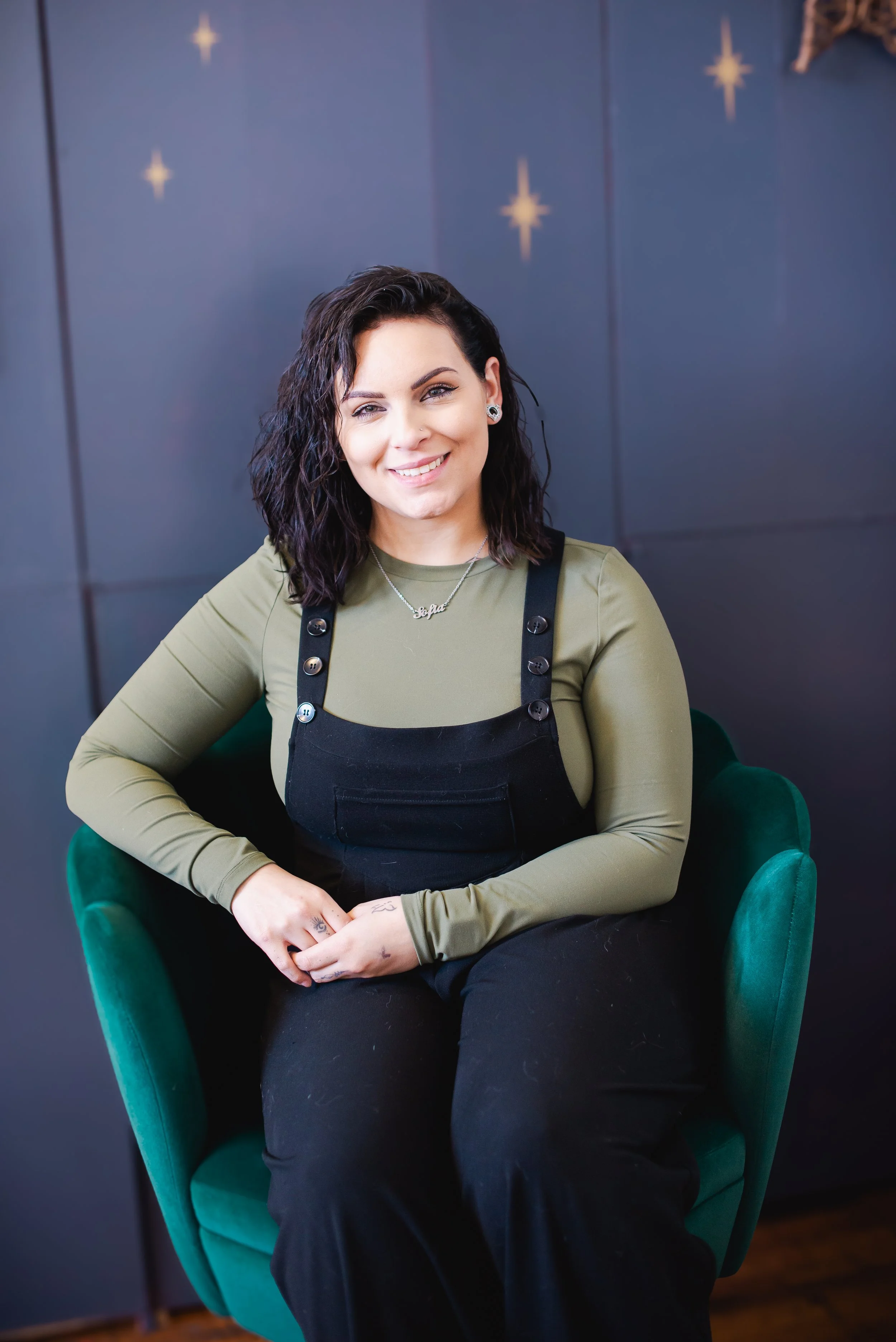 A woman with short dark wavy hair, wearing an olive green long sleeve shirt and black overalls, sitting on a green armchair with a dark background decorated with stars, smiling at the camera.