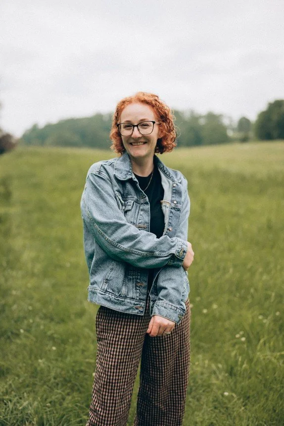 Smiling woman with short curly red hair and glasses, wearing a denim jacket and patterned pants, stands in a green field. EMDR & Trauma Therapists in CT.