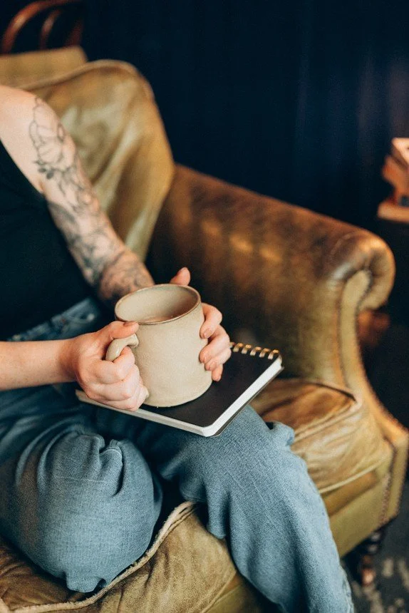 Person seated in a leather armchair with their face out of view wearing jeans and a black top which shows a tattooed arm. The person is holding a cream colored coffee mug with both hands and underneath the mug rests a black notebook.