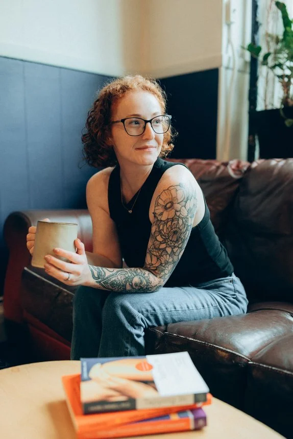 Curly red hair woman with glasses and arm tattoos sits on leather couch holding mug near EMDR books. Professional CT EMDR & trauma therapists guiding curious, overwhelmed minds.