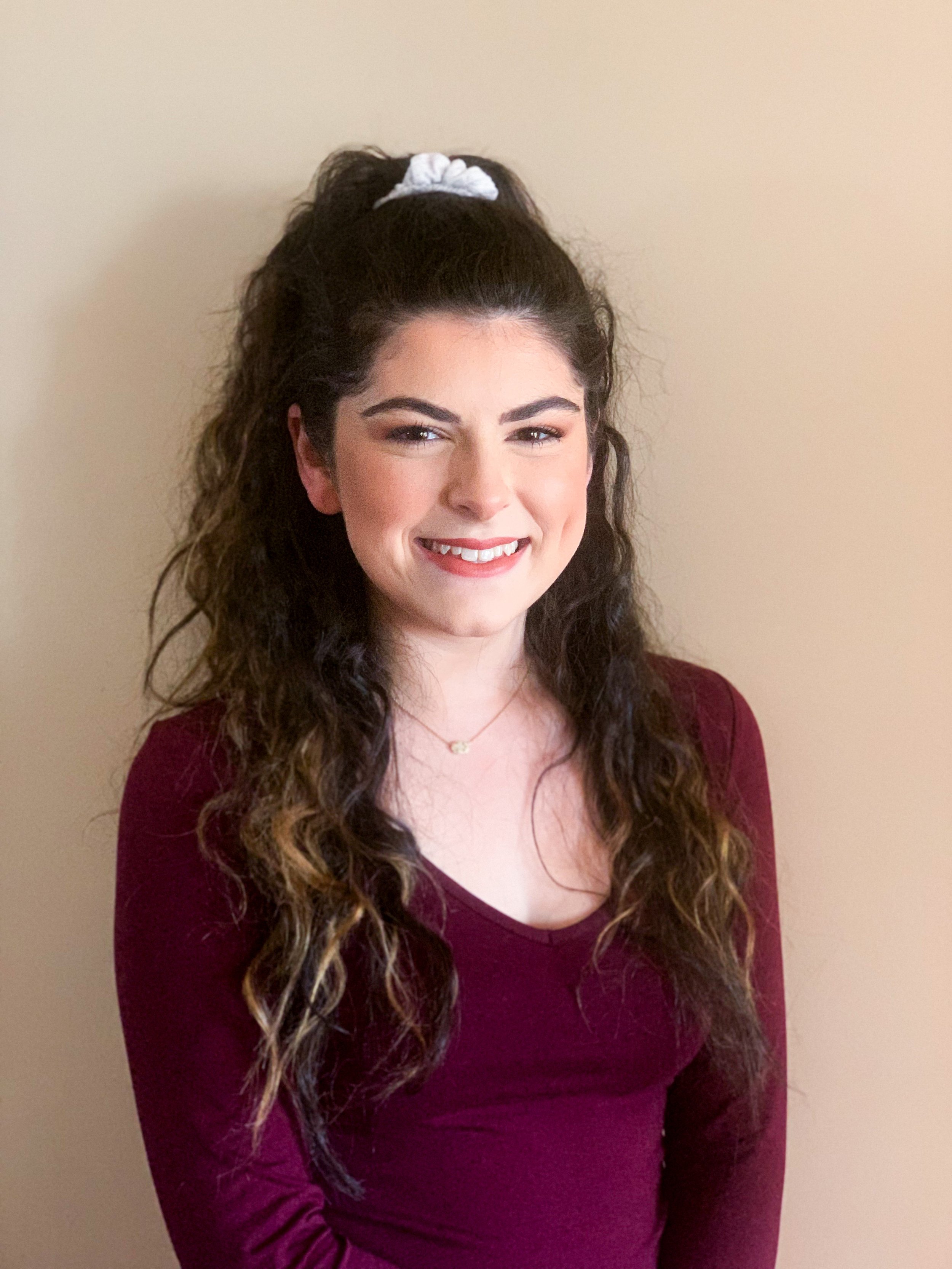 Smiling woman with long, curly dark hair tied back with a white scrunchie, wearing a maroon long sleeve shirt and a thin necklace against a neutral wall. Dedicated young adult therapist.