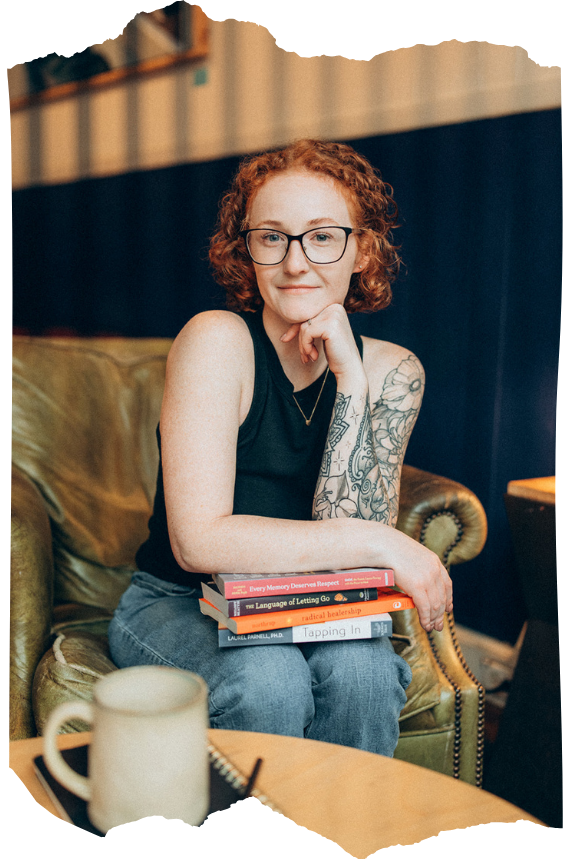 Woman with short curly red hair and black-framed glasses sits on a leather armchair holding a stack of books, featuring intricate arm tattoos. Professional EMDR and trauma therapist in CT.