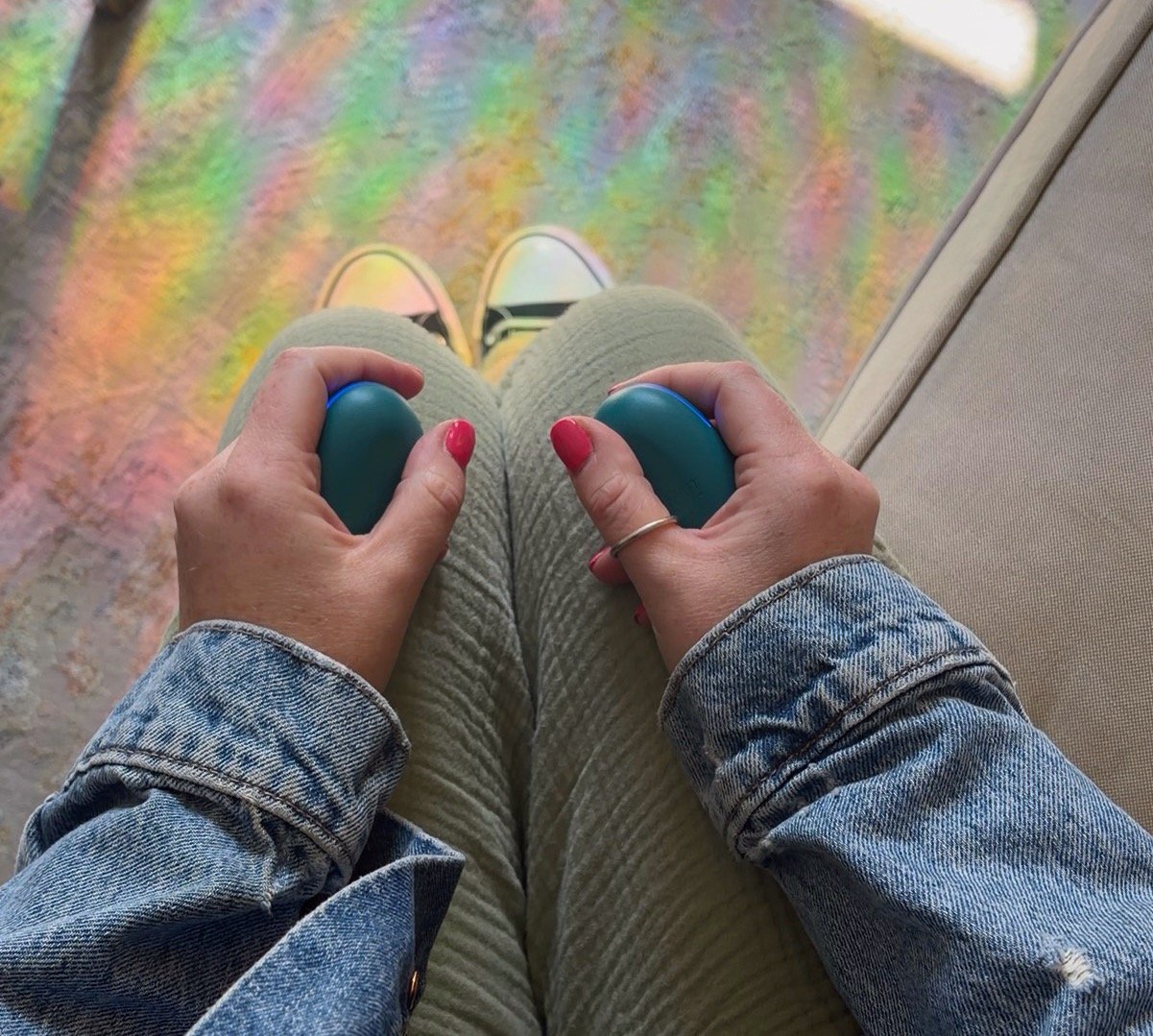 Hands with pink nails holding blue EMDR buzzers during a bilateral stimulation session over olive pants and a rainbow tinted rug. Experiencing specialized EMDR therapy in Connecticut.