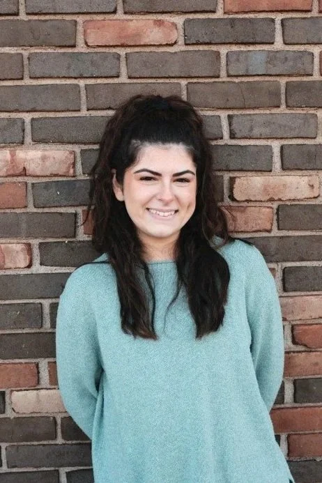 Smiling young woman with long dark hair, wearing a teal sweater, leans against a brick wall. Representing compassion and care as part of a team of EMDR & Trauma Therapists in CT.