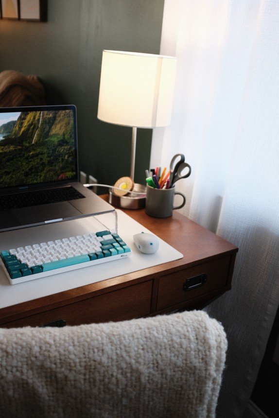 Laptop on clear stand with teal keyboard, computer mouse, and glowing lamp on wooden desk by white curtain. Cozy remote workspace optimized for virtual EMDR consultation with EMDR Approved Consultant.