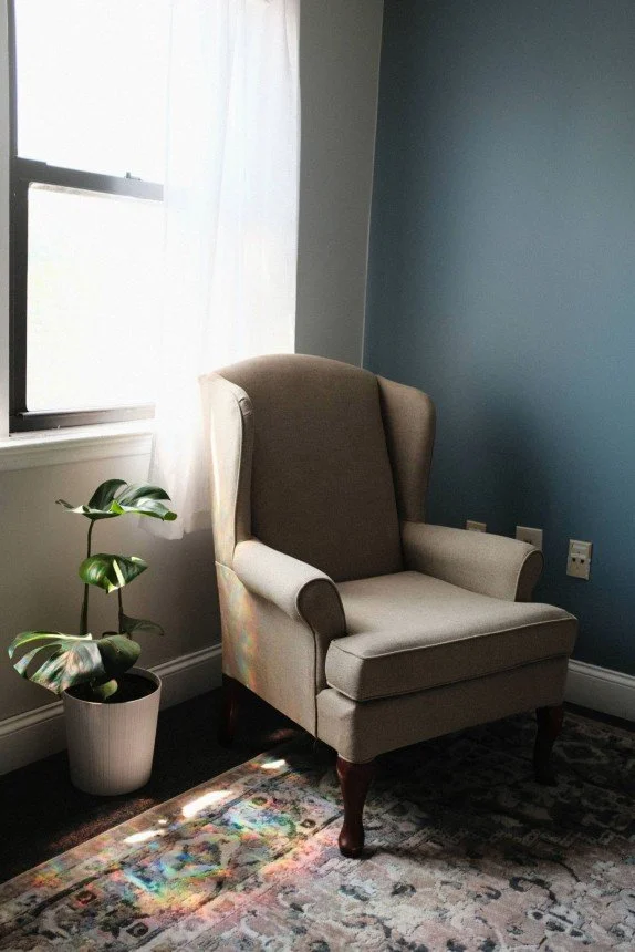 Green armchair in front of a brightly lit window with a blue wall on one side and a small monstera plant on the other, depicting a cozy therapy office