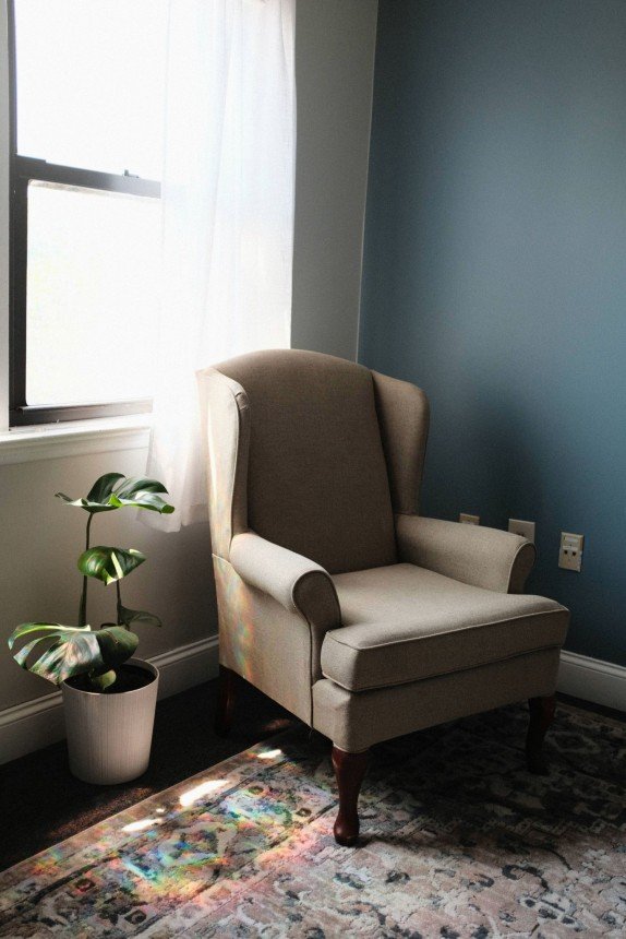 Beige wingback armchair sits by a window with white curtains, a potted monstera plant, and a blue wall. Safe space offering therapy for overwhelmed young adults.