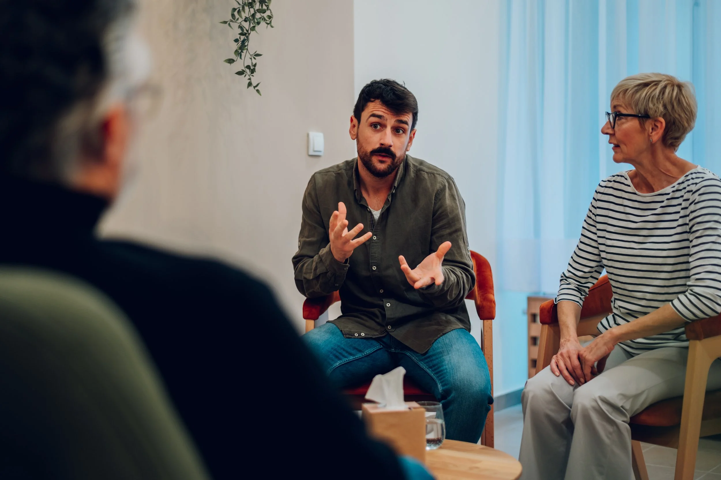 A man with dark hair and a beard speaking to a group in a casual setting. The image also shows an older woman with short blonde hair and glasses, sitting to his right, listening. Another person is partially visible in the foreground.