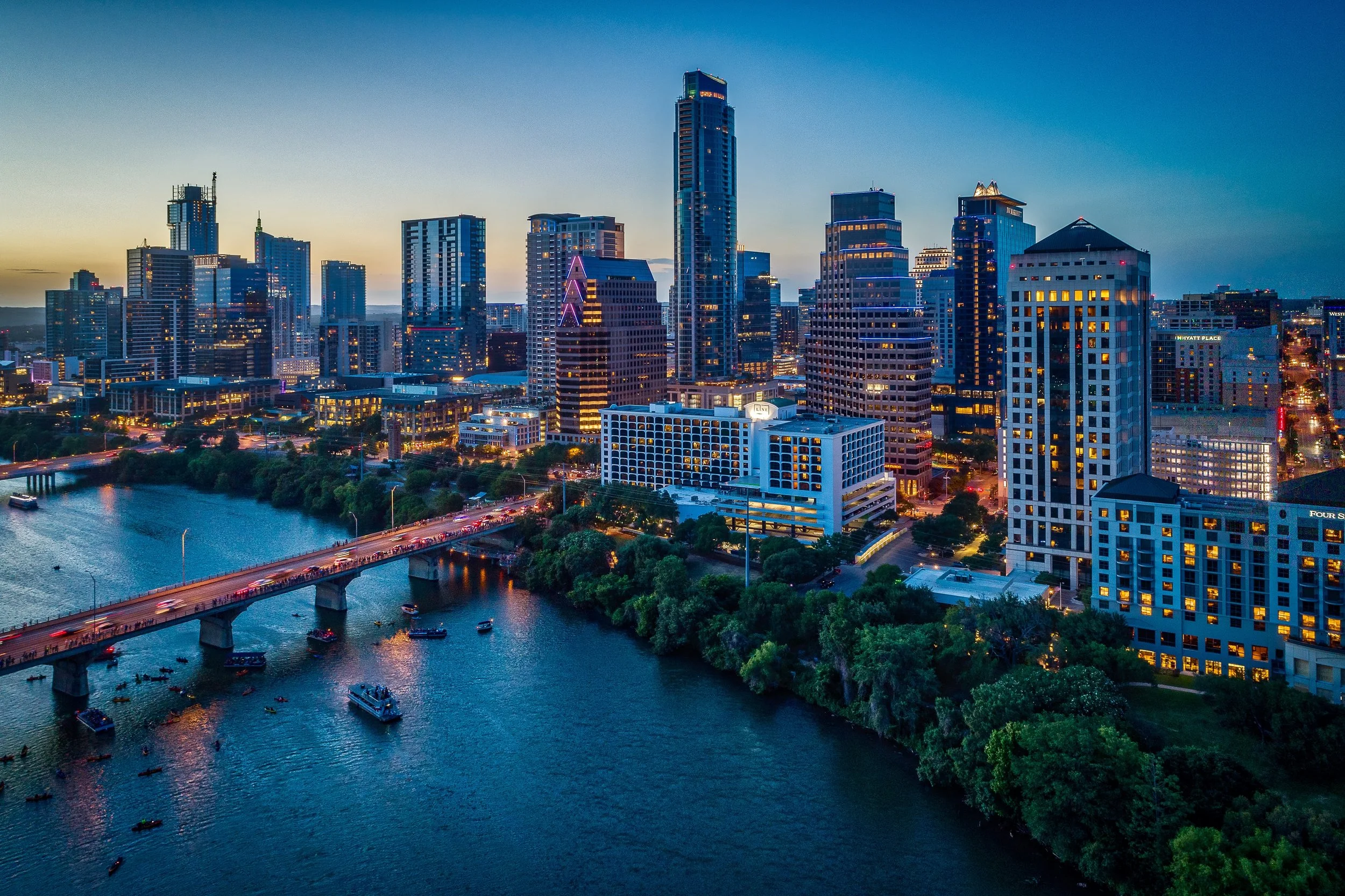 An aerial view of a city skyline at dusk, featuring tall modern skyscrapers, a river, and a bridge with boat traffic in the foreground.
