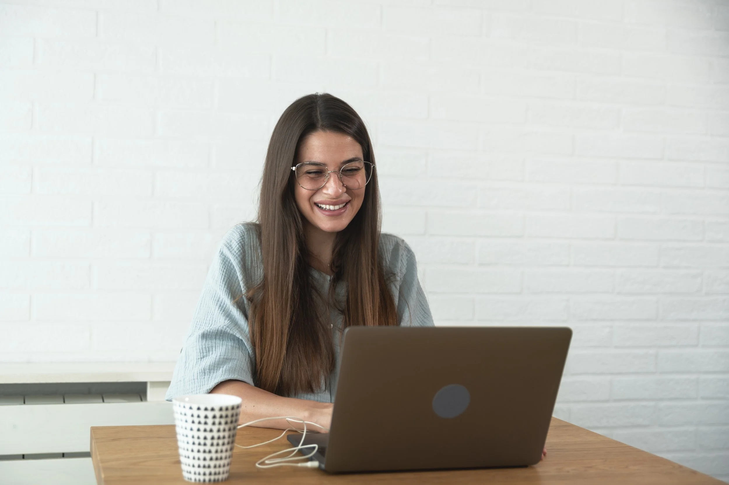Young woman with long brown hair and glasses smiling while sitting at a wooden table, using a laptop.