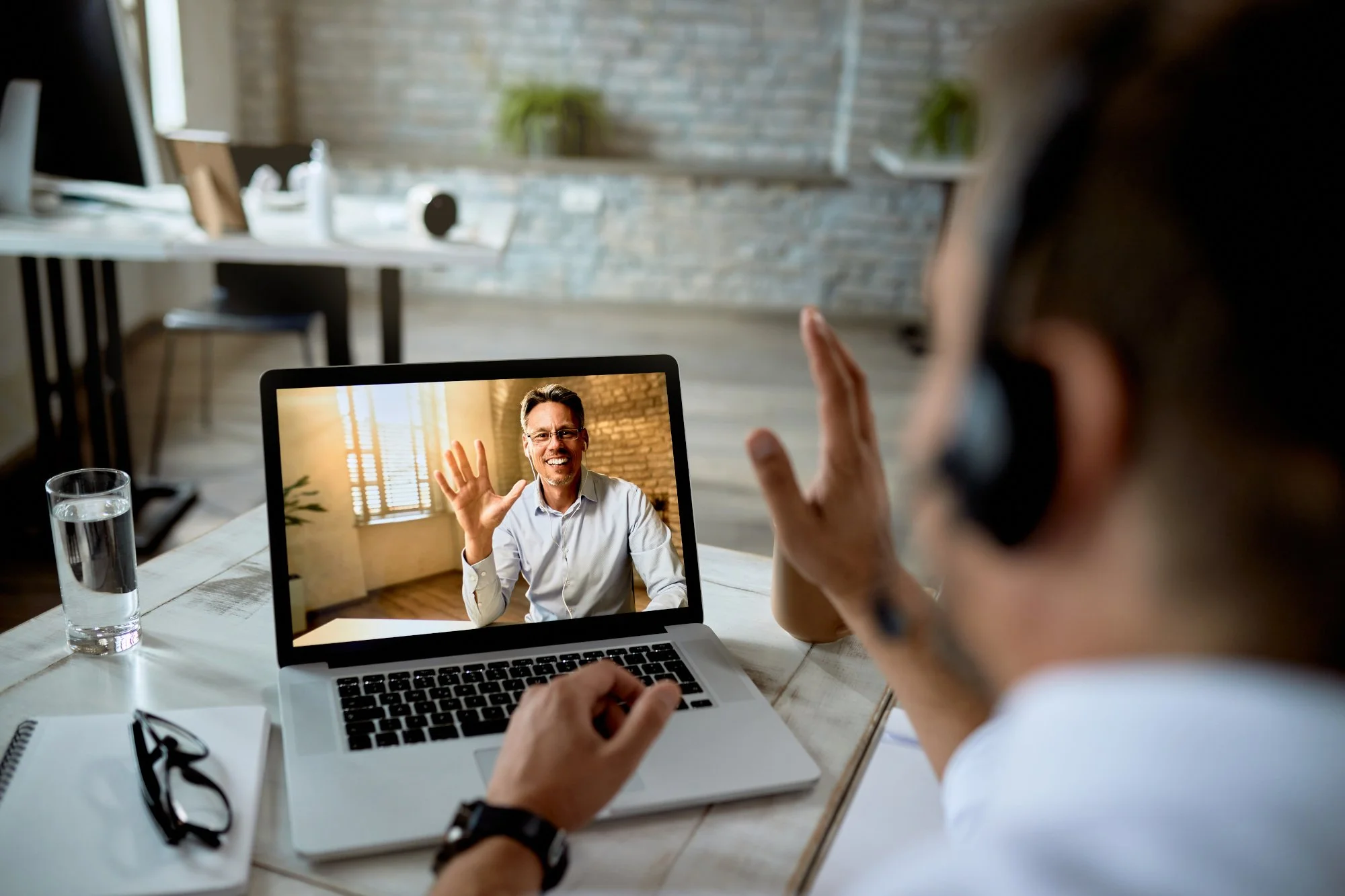 Person on a video call waving to another person on a laptop screen, in an office setting with a glass of water, glasses, and notebooks on the desk.