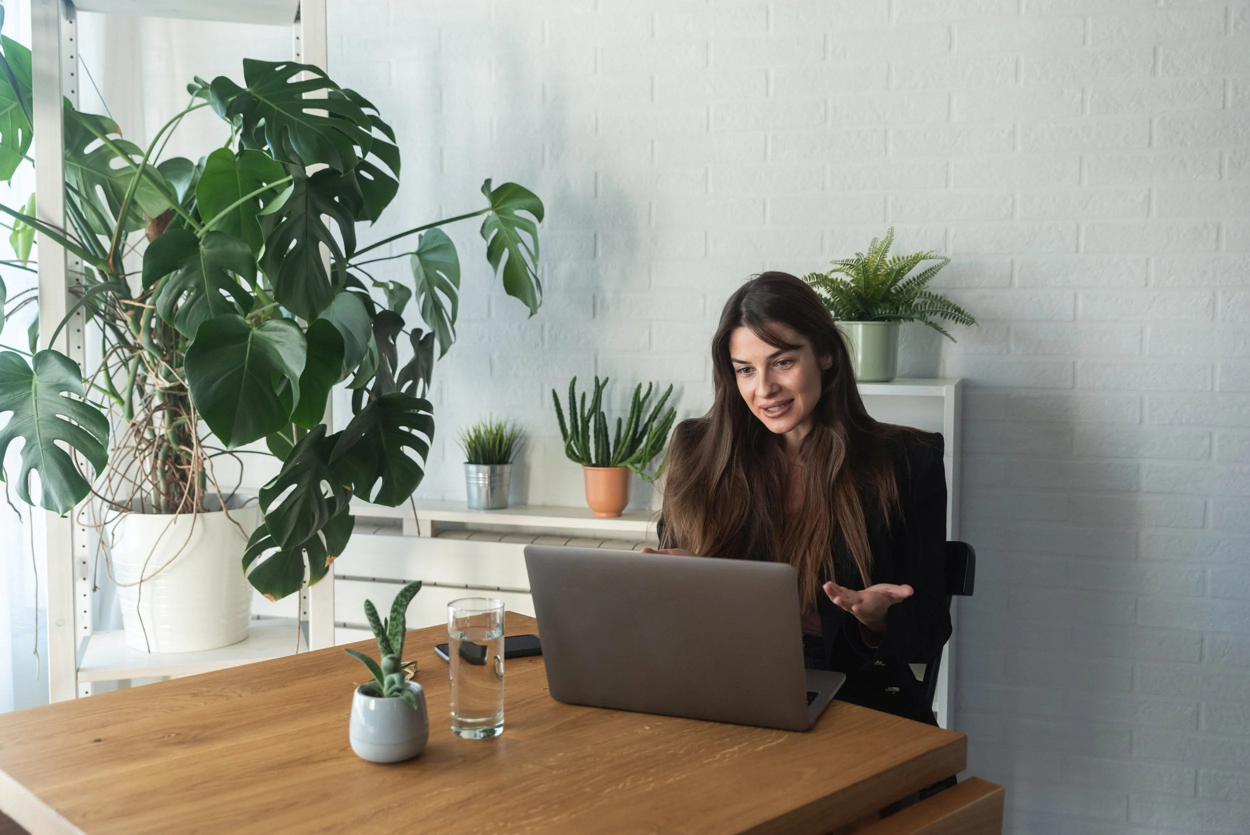 A woman with long brown hair working on a laptop at a wooden desk in a bright room with white brick walls and various potted plants, including a large monstera, on shelves and the desk.