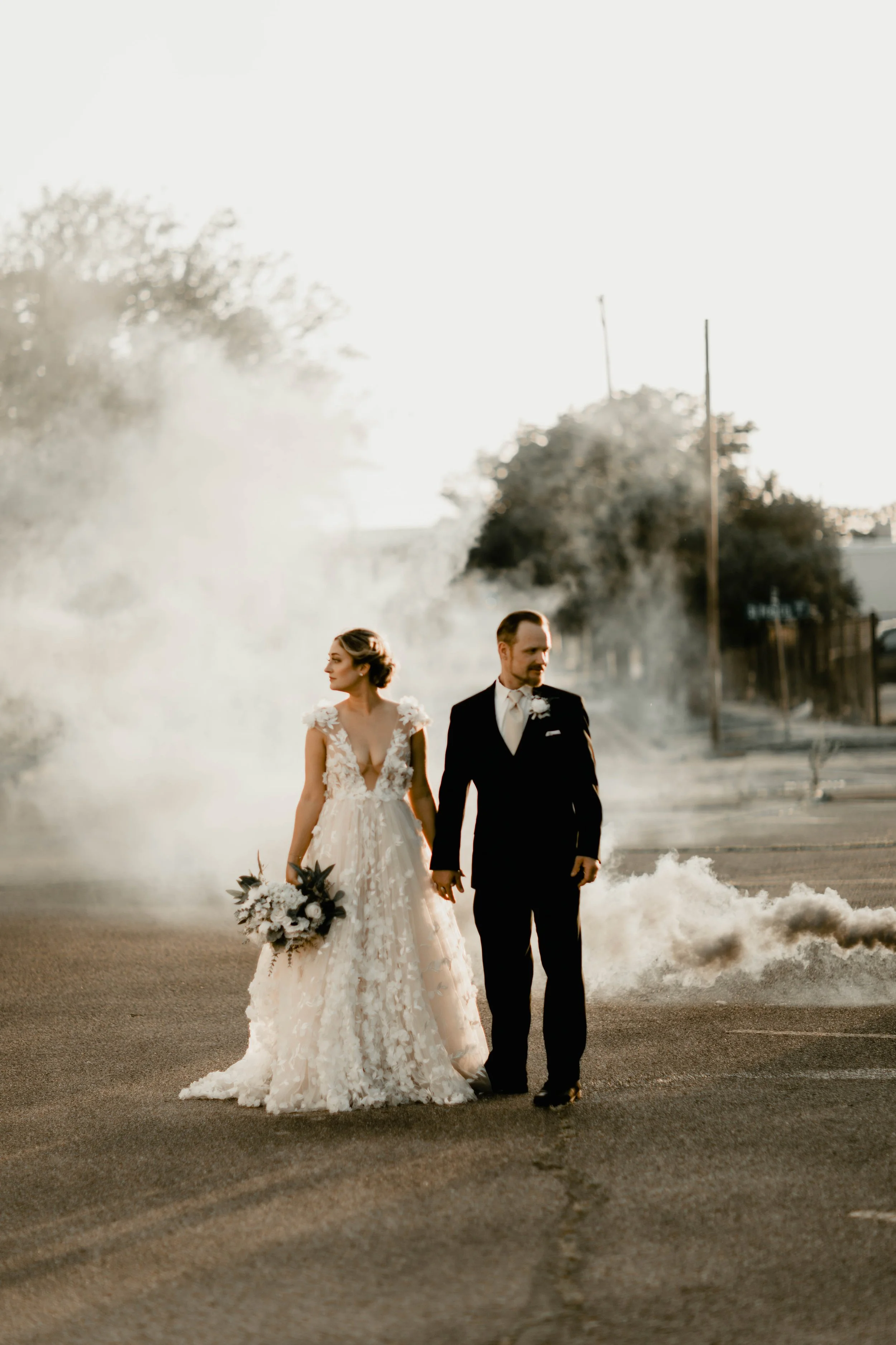 Bride and groom holding hands walking in an empty parking lot with smoke and backlit trees in the background.