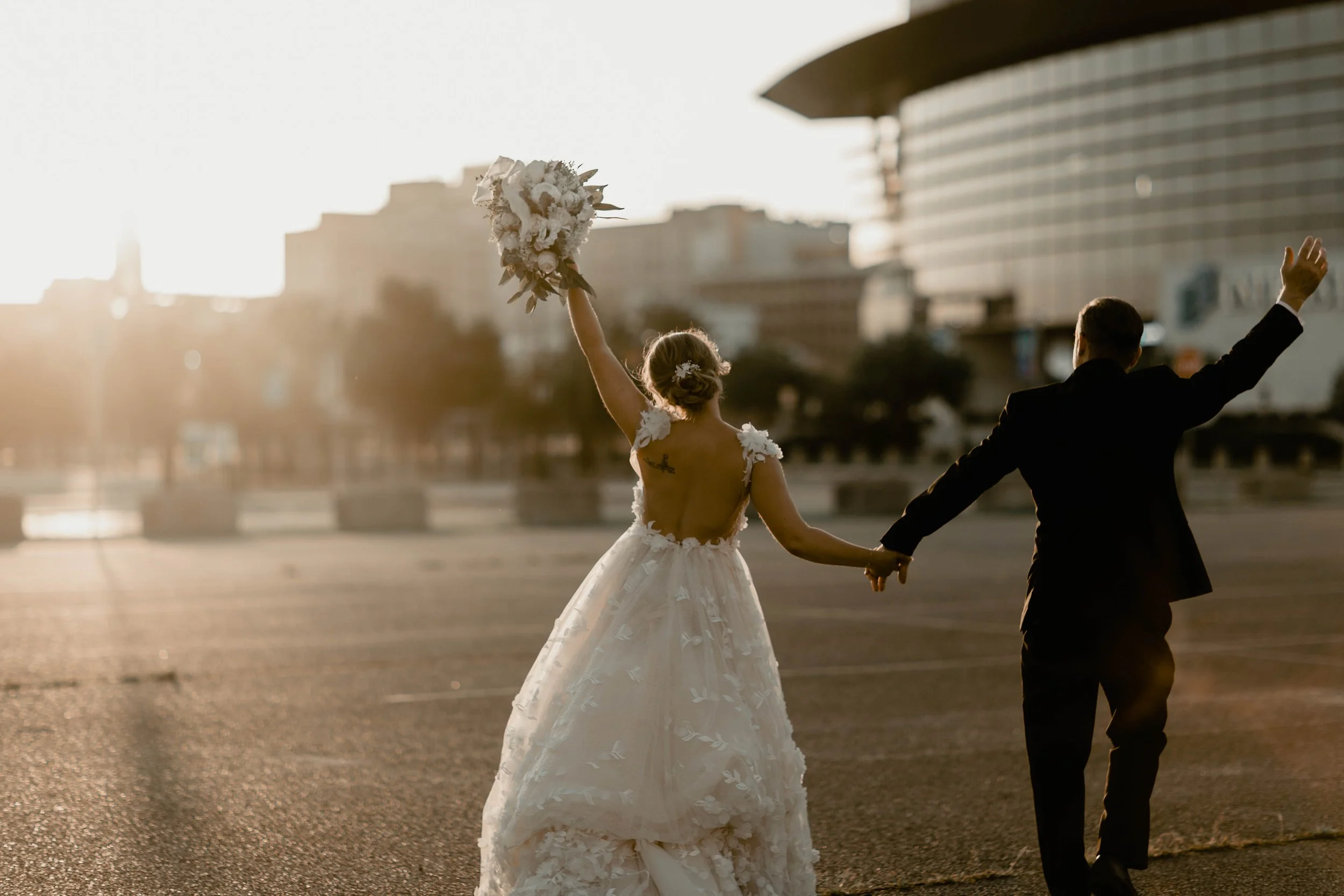 A bride and groom walking hand in hand in an urban area during sunset, with the bride holding her bouquet up and wearing a wedding dress, the groom in a black tuxedo.