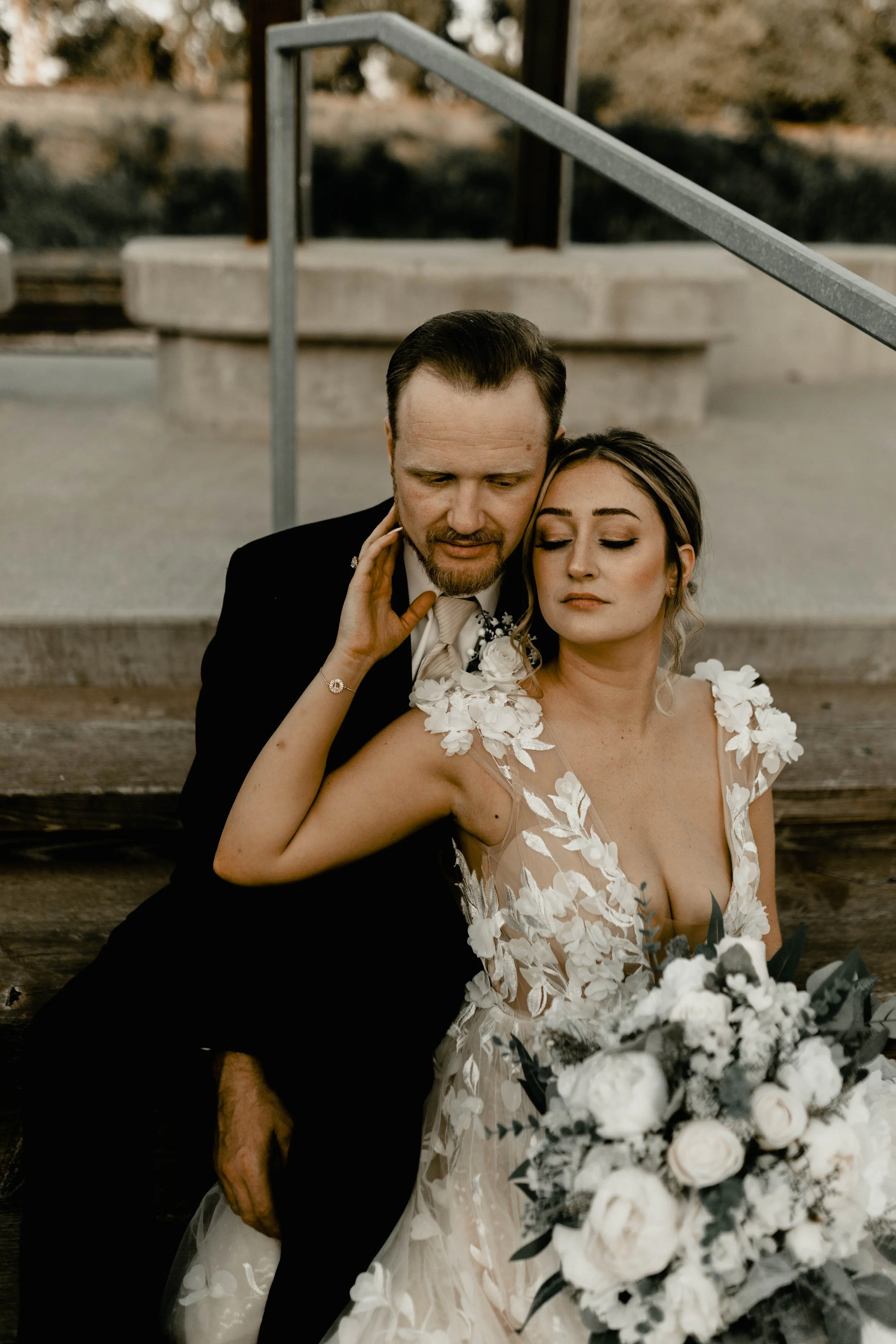 A bride and groom sitting closely with eyes closed; the bride is in a wedding dress holding a bouquet, the groom in a black suit, outdoors in a natural setting.