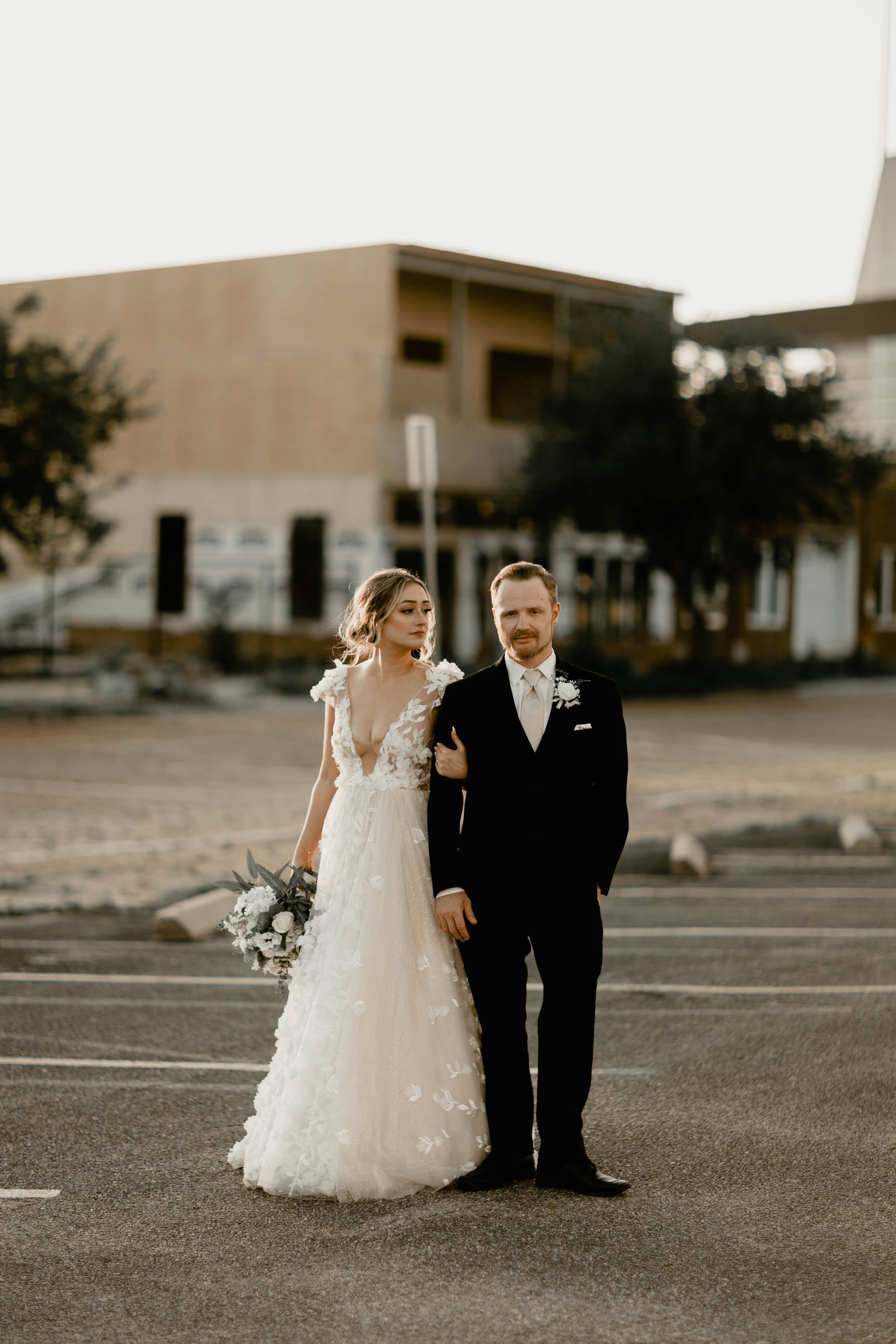 A bride in a white wedding gown holding a bouquet stands arm-in-arm with a groom in a black tuxedo outside in a parking lot at sunset.