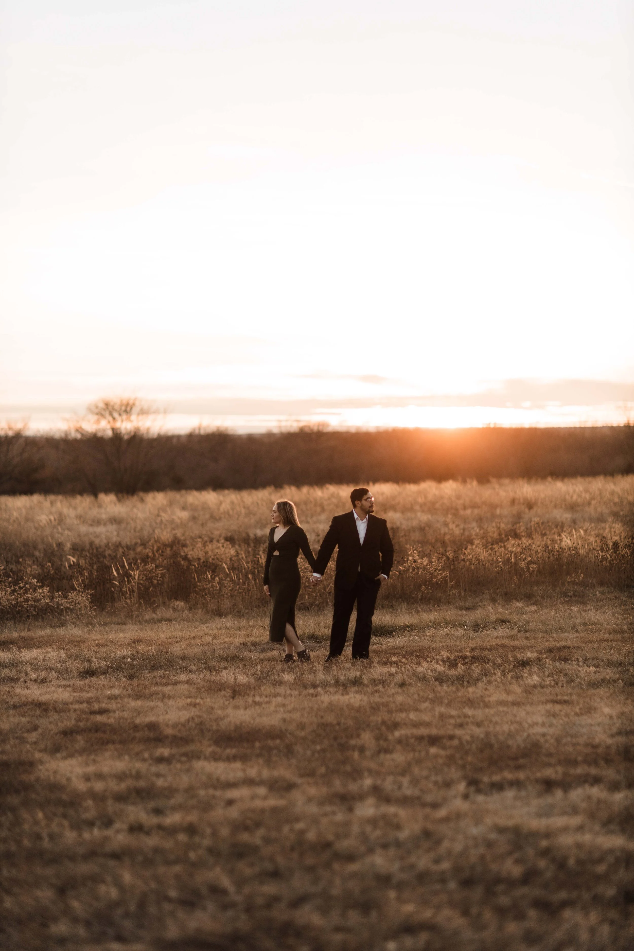 A man and woman in formal attire walking hand in hand through an open field at sunset.