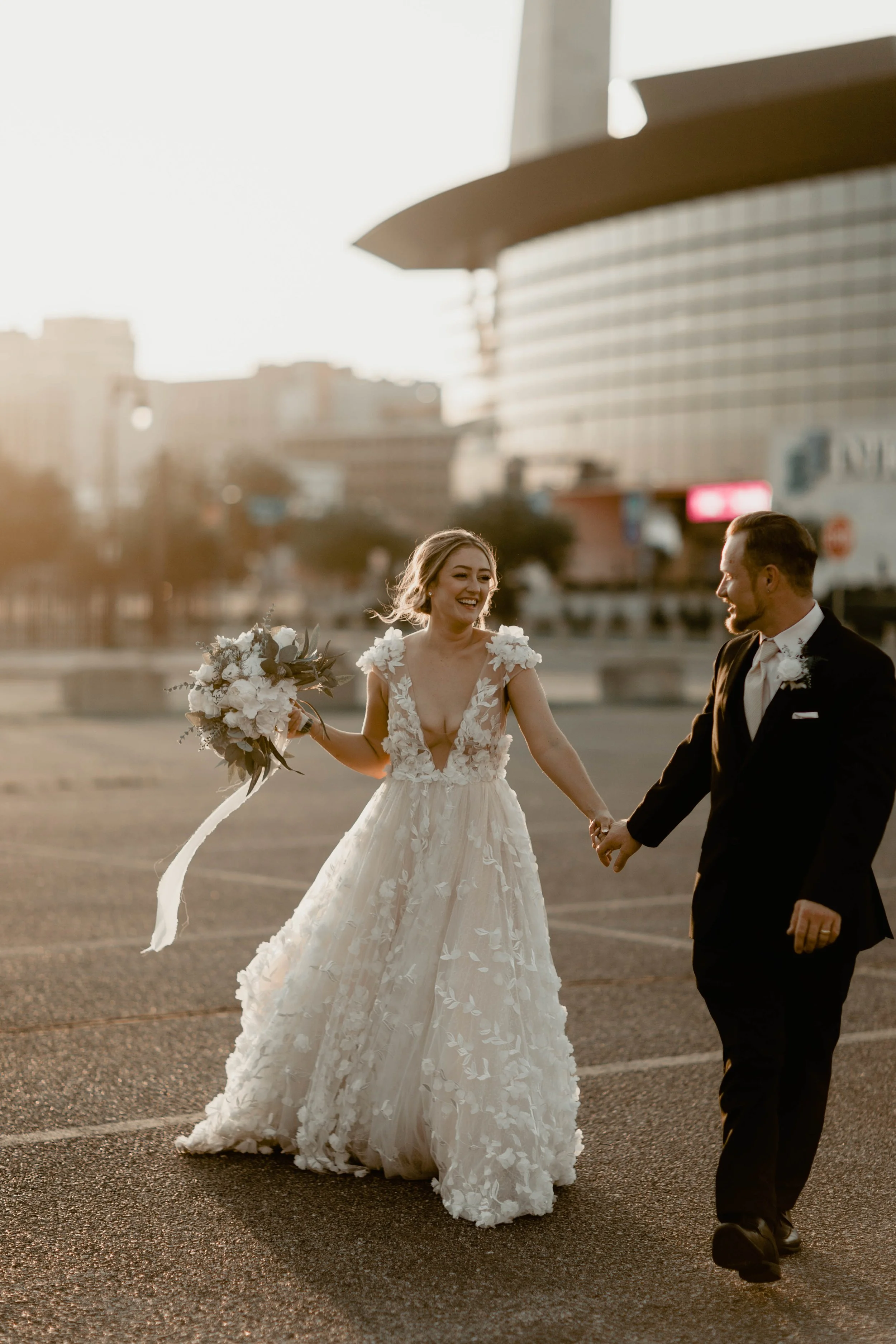 A bride and groom holding hands and smiling, with the bride in a white wedding dress holding a bouquet, in an outdoor urban setting during sunset.