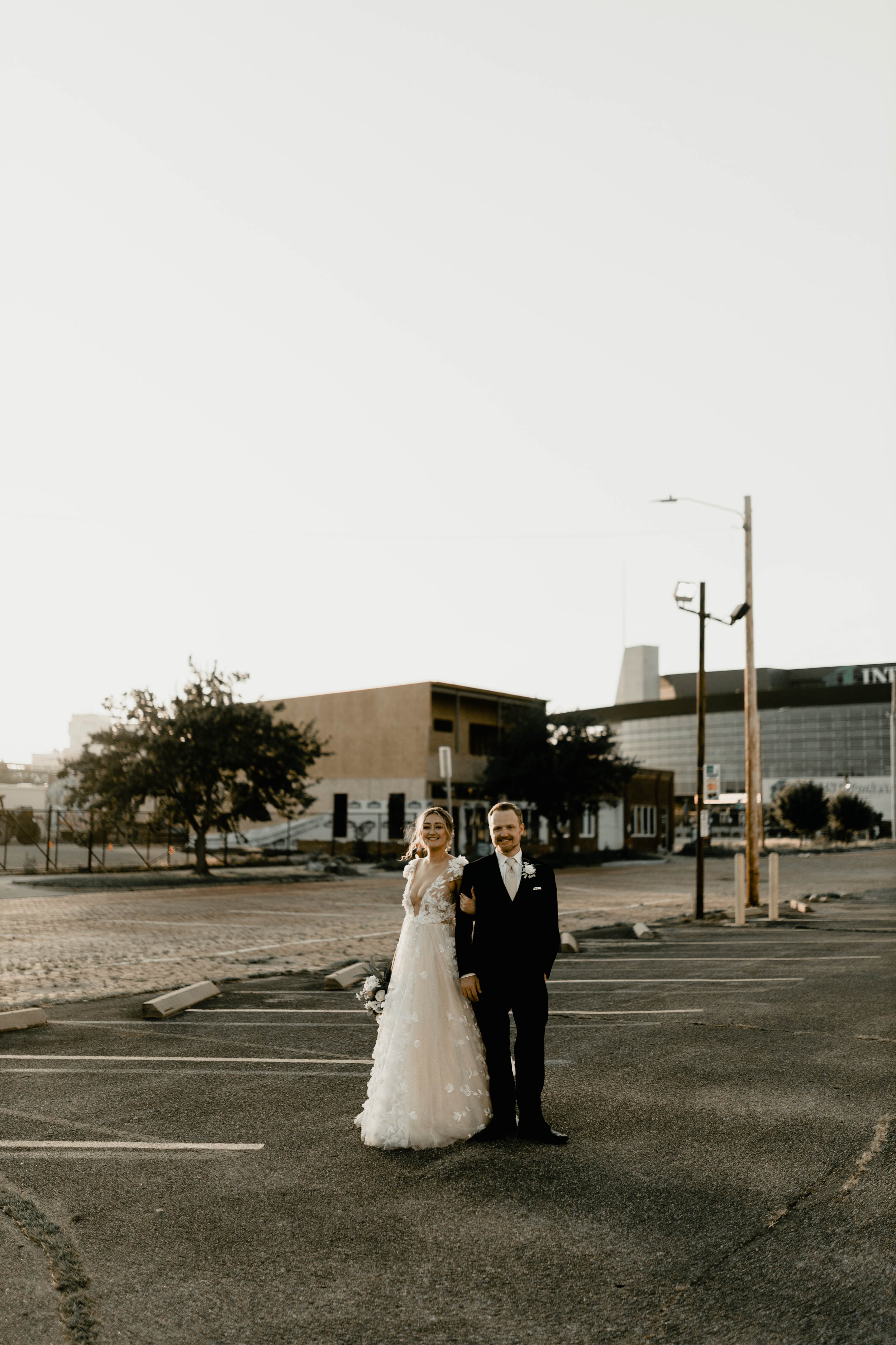 A newlywed couple stands in an empty parking lot during sunset, smiling and dressed in wedding attire with a cityscape background.