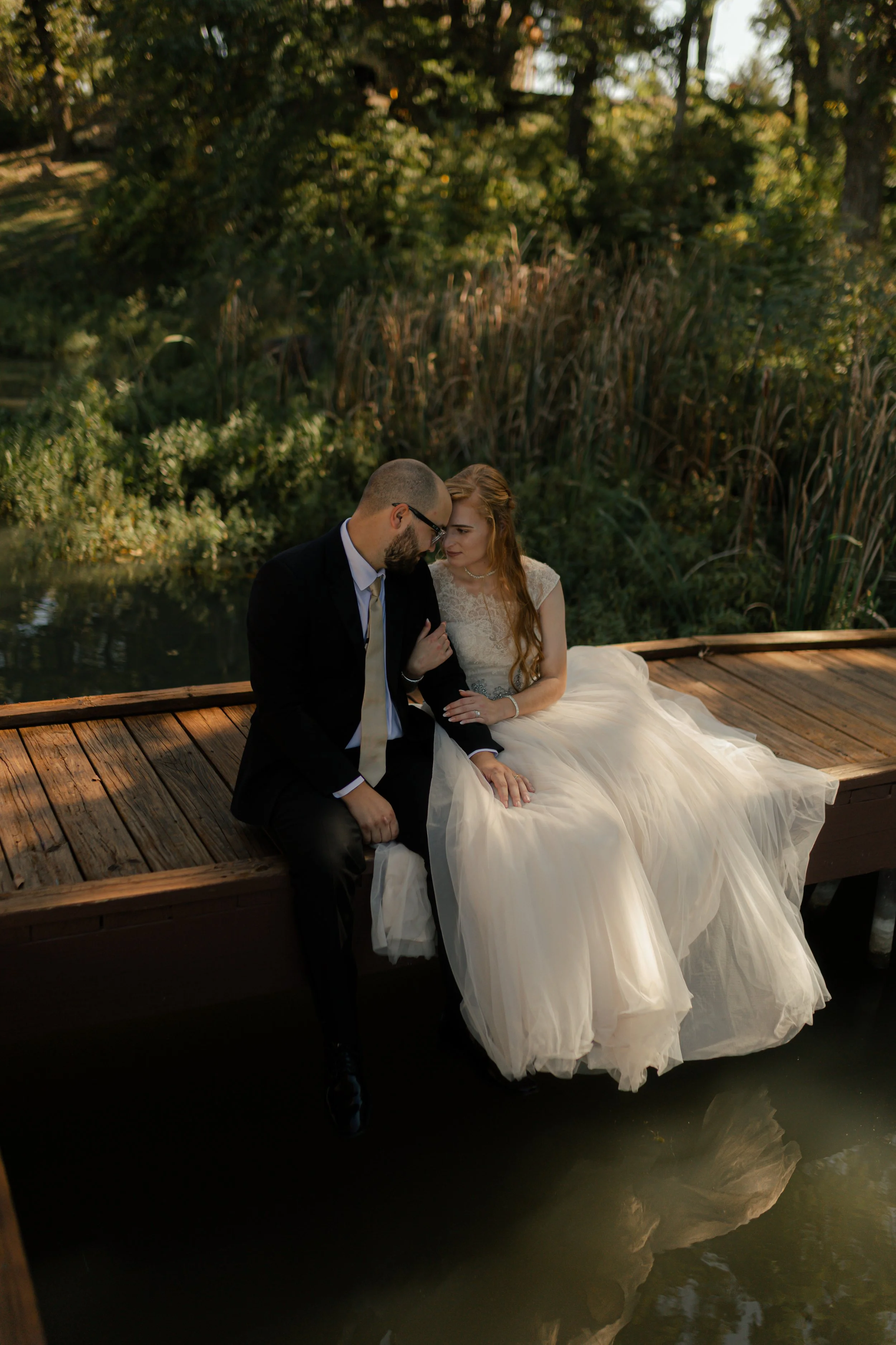 A newlywed couple sitting on a wooden dock by a pond, sharing an intimate moment, with trees and tall grass in the background.