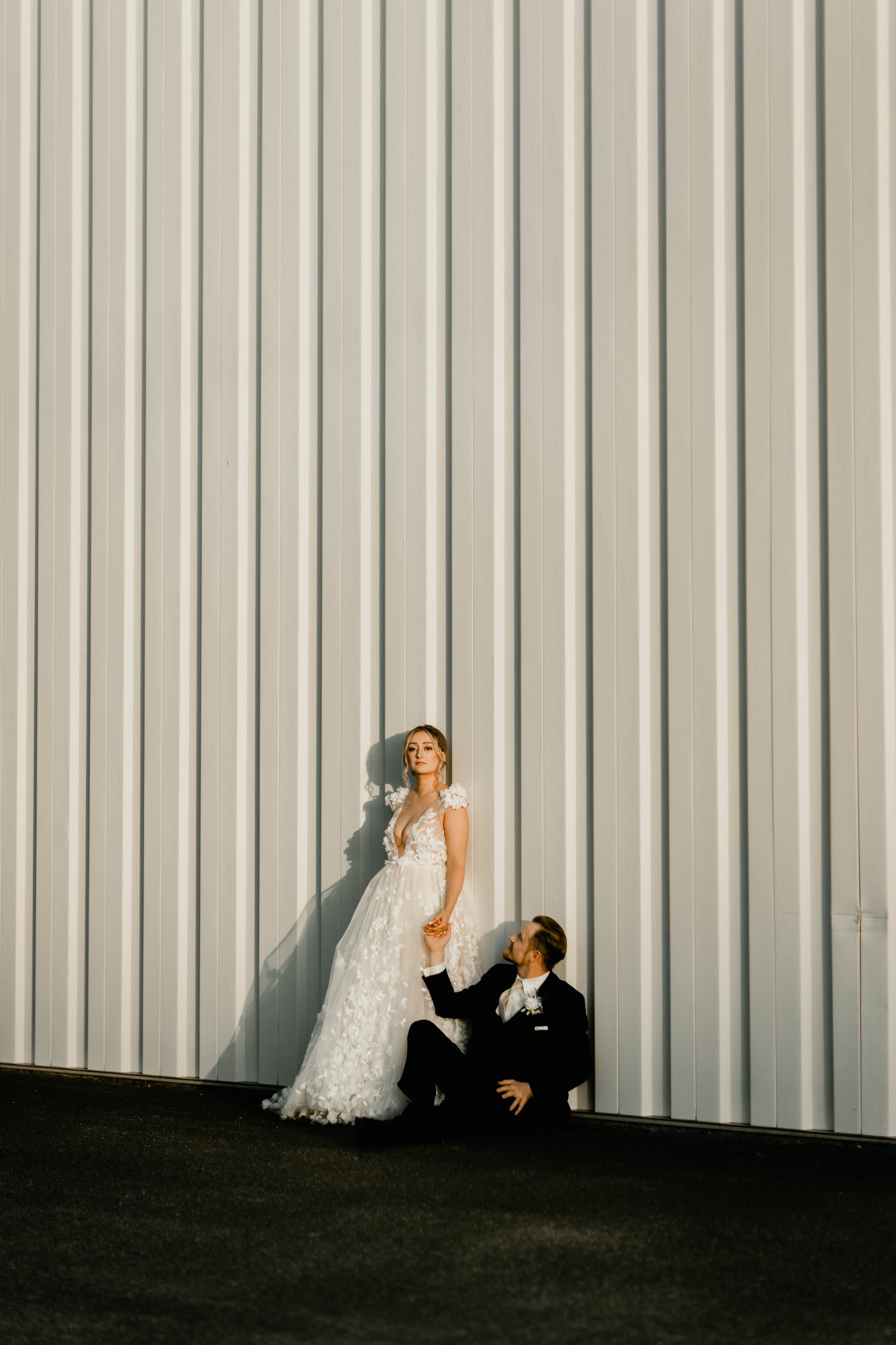 A bride and groom in wedding attire, standing and sitting against a large, white, corrugated metal wall during sunset.