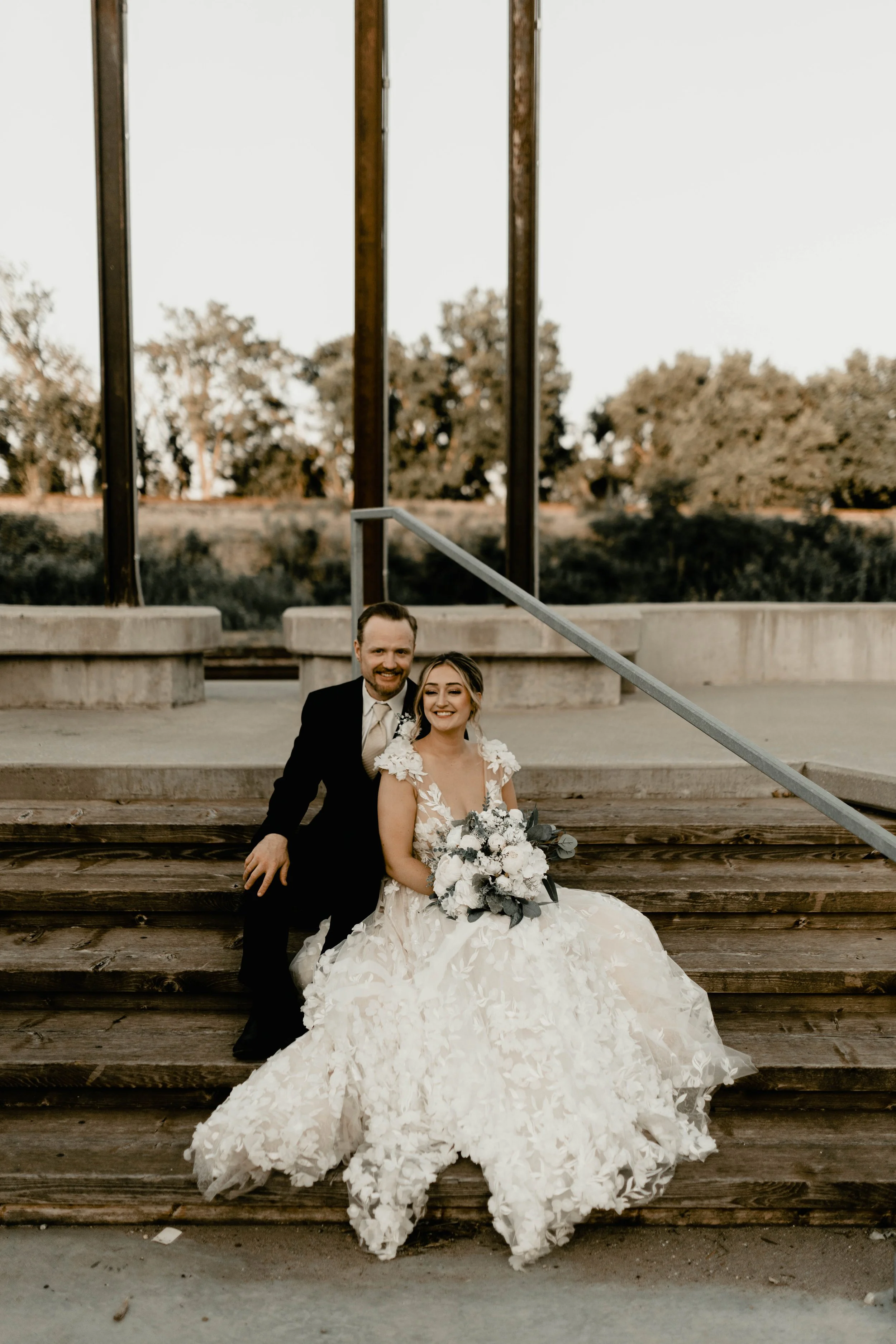 A bride in a white wedding dress holding a bouquet, sitting on wooden stairs next to a groom in a black suit outdoors at sunset.