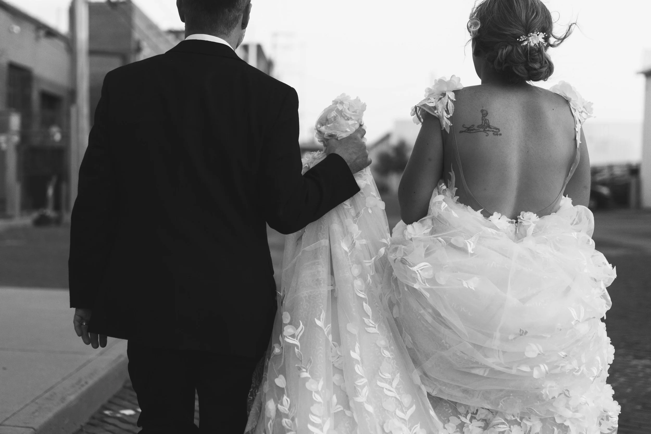 A couple dressed in wedding attire walking away on a city street, with the woman holding her wedding gown.