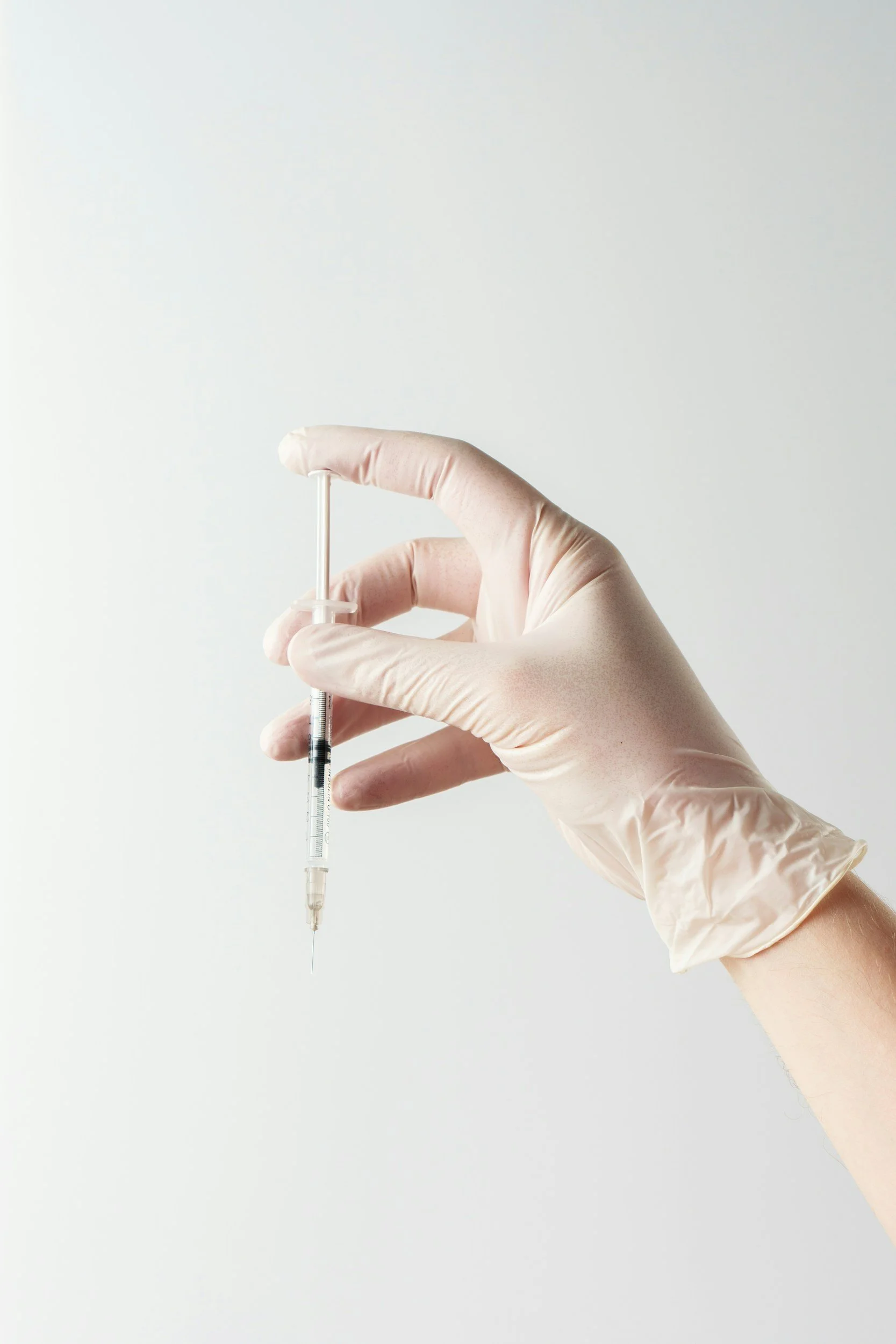 Hand wearing a latex glove holding a syringe with a needle against a plain white background.