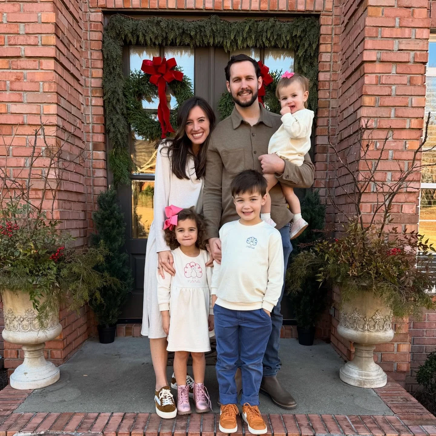 Family of five standing on front porch decorated with Christmas wreath, plants, and brick walls.